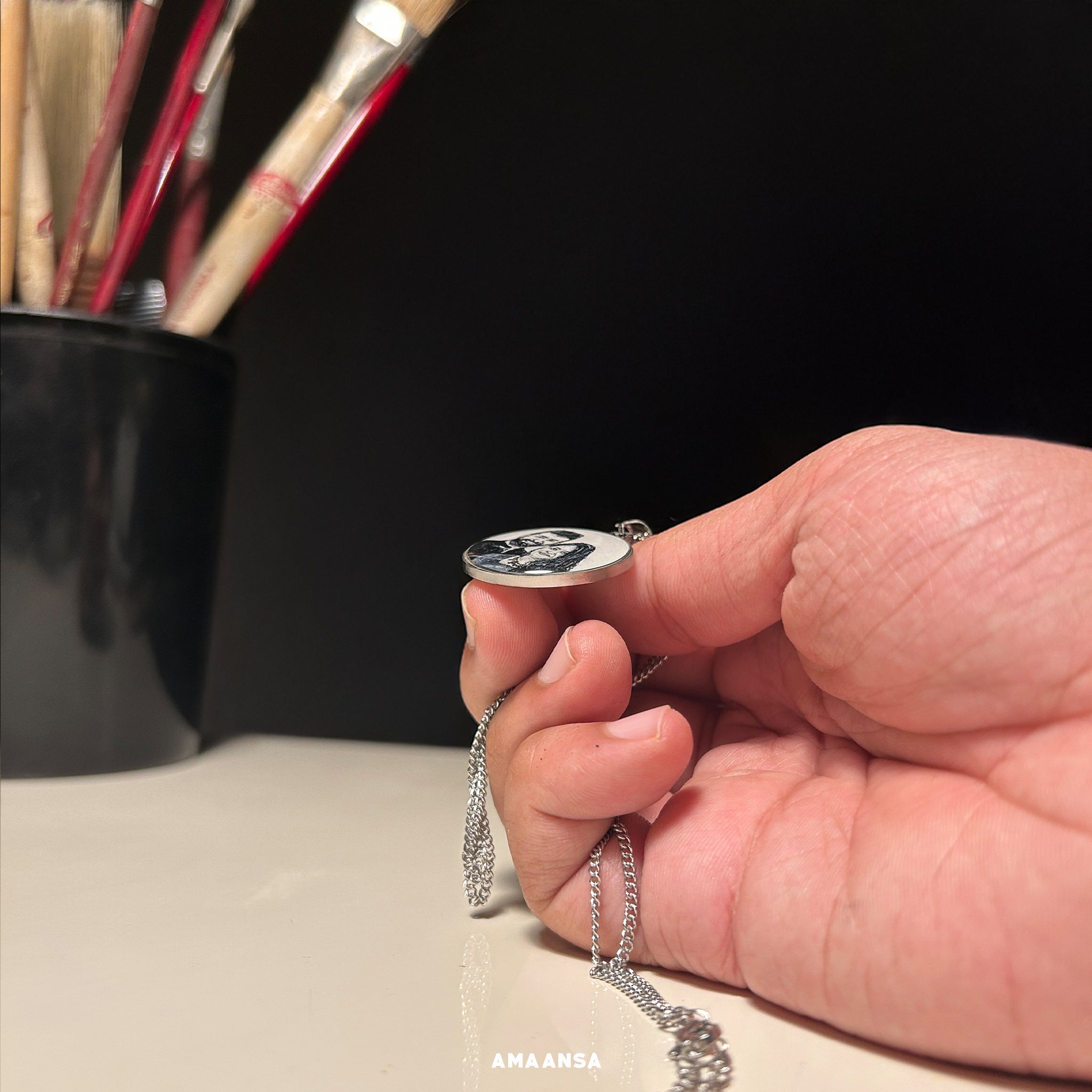 "Artisan perspective of AMAANSA's 26mm pendant - showcasing the refined profile of rhodium-colored miniature portrait, delicately balanced between thumb and forefinger to demonstrate its sleek depth and premium finish, chain gracefully cascading against studio backdrop with artist's brushes in soft focus, emphasizing the pendant's professional craftsmanship and elegant proportions