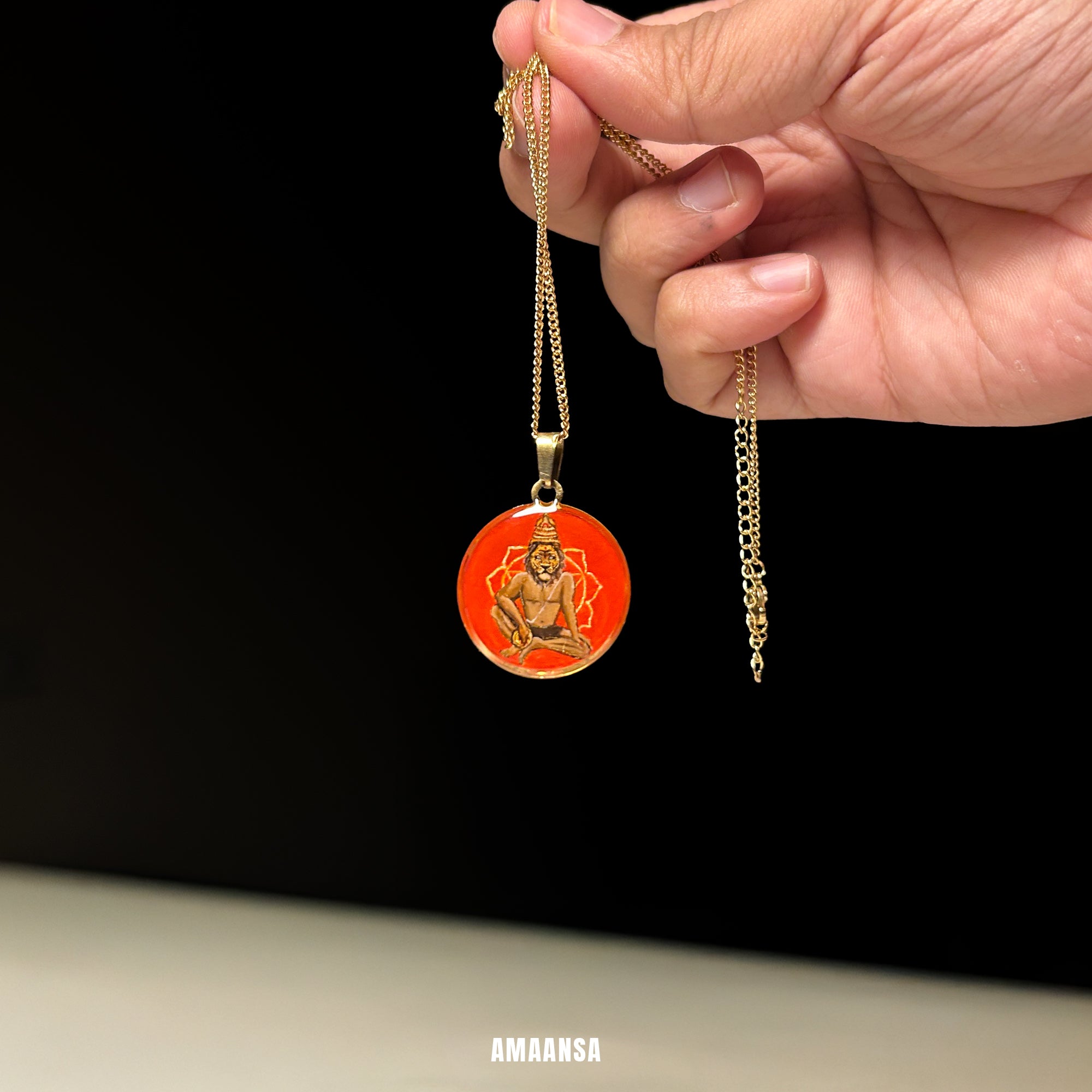 Hand holding a gold necklace with an red pendant featuring a deity on a black background. Custom deity locket handpainted Lord Narasimha full color miniature painting red background gold foil paper 18k gold PVD pendant stainless steel chain devotional jewelry protection talisman spiritual keepsake studio palette backdrop