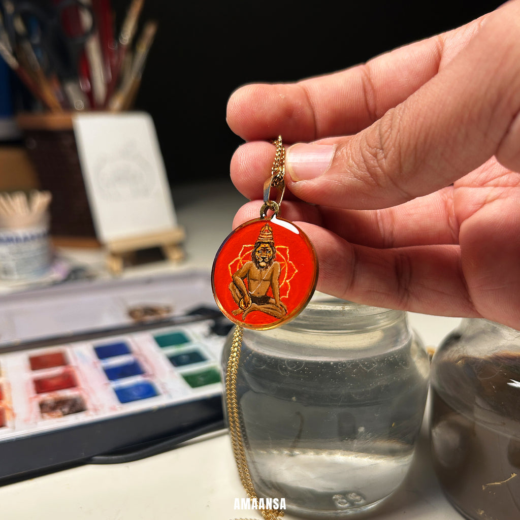 Hand holding a red pendant with a gold chain in front of art supplies. Handpainted Narasimha avatar locket full color miniature art red background gold foil 18k gold PVD pendant stainless steel chain divine protection religious jewelry devotional talisman artist studio setting