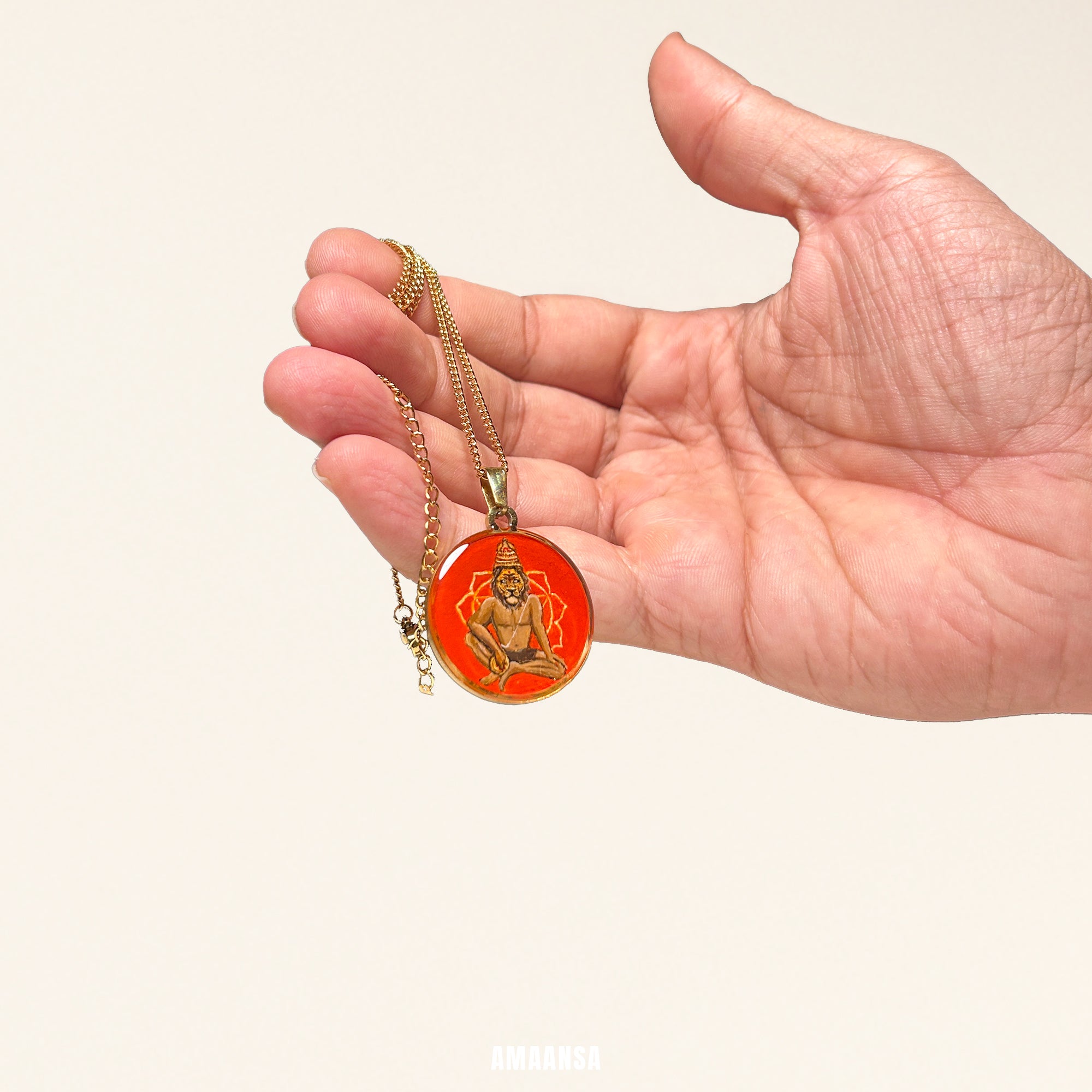 Hand holding a gold necklace with an red pendant featuring a seated figure on a beige background. 18k gold PVD locket custom Lord Narasimha miniature painting vibrant red background gold paper devotional coin spiritual protection jewelry religious necklace professional studio shoot


