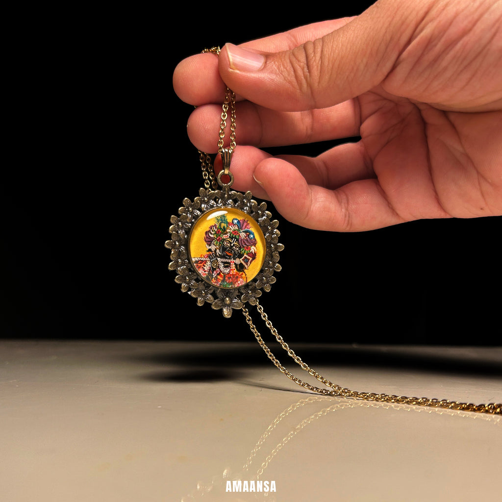 Hand holding a decorative necklace with a  pendant against a black background. Custom deity locket handpainted Banke Bihari Lord Krishna semicolor miniature painting devotional jewelry antique metal patina chain vintage yellow background Vrindavan Krishna divine protection religious keepsake