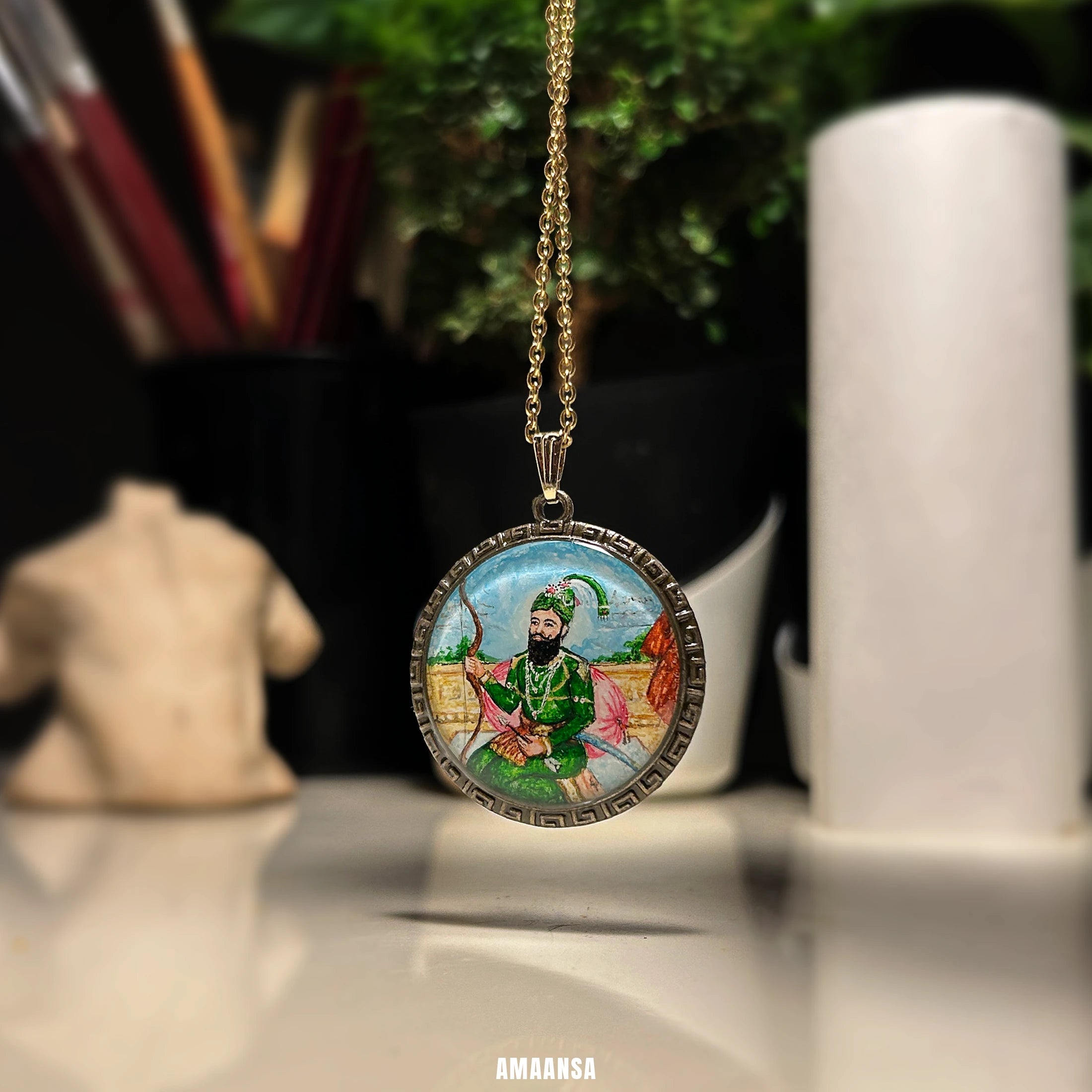 Round Guru Gobind Singh miniature portrait pendant on chain, detailed painting framed by antique brass‑tone border against white cylinder.
