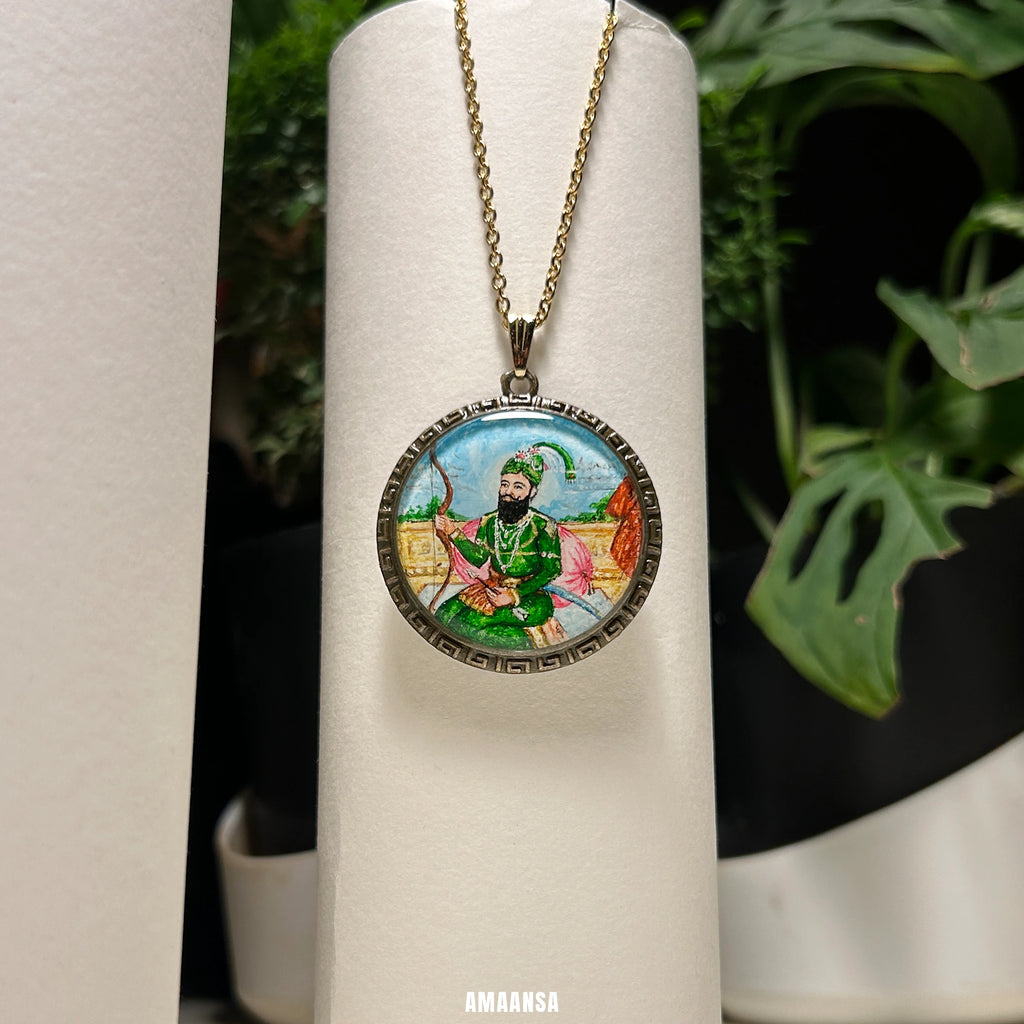 Hand‑painted Guru Gobind Singh miniature portrait pendant hanging on chain, devotional necklace styled among art brushes and plants.