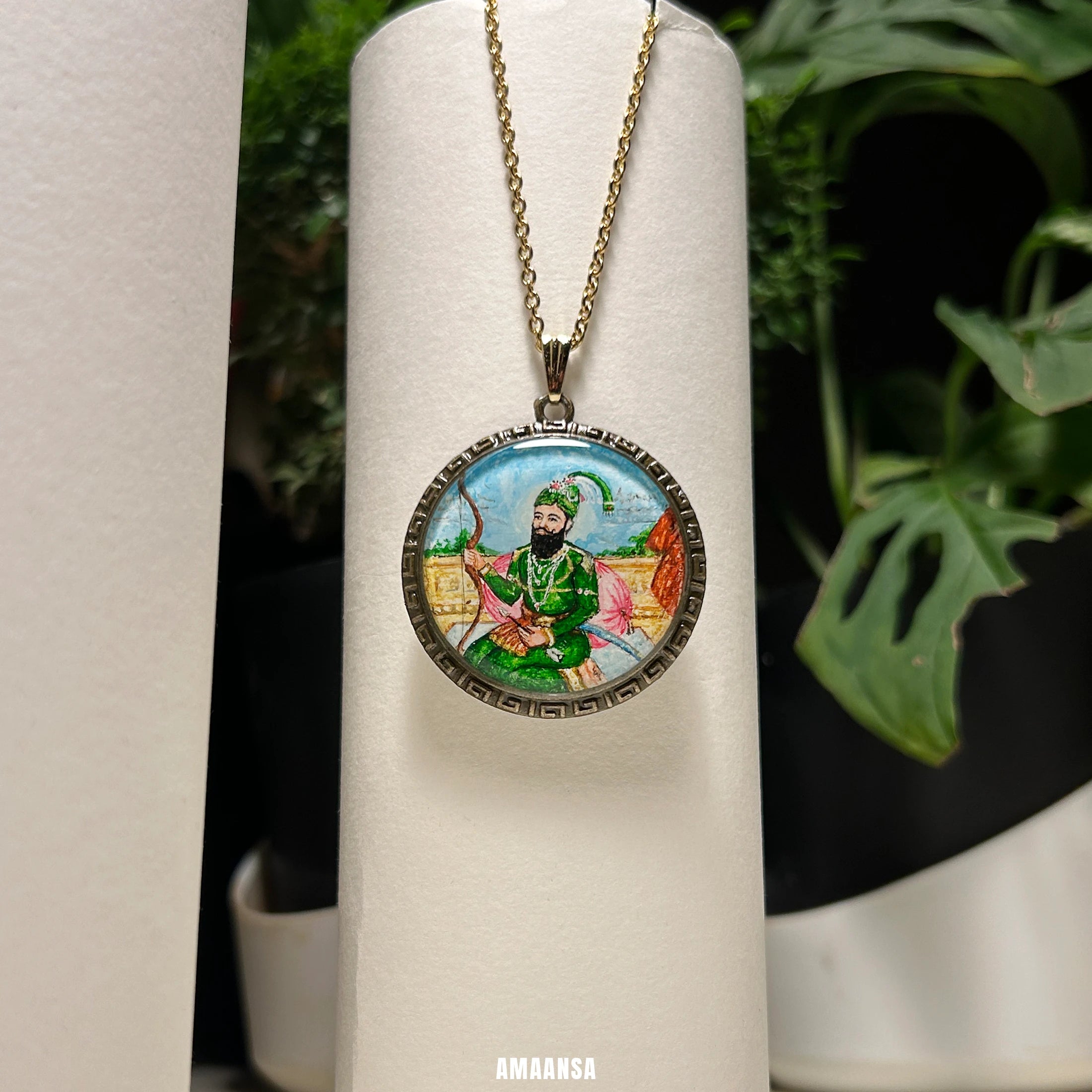 Hand‑painted Guru Gobind Singh miniature portrait pendant hanging on chain, devotional necklace styled among art brushes and plants.