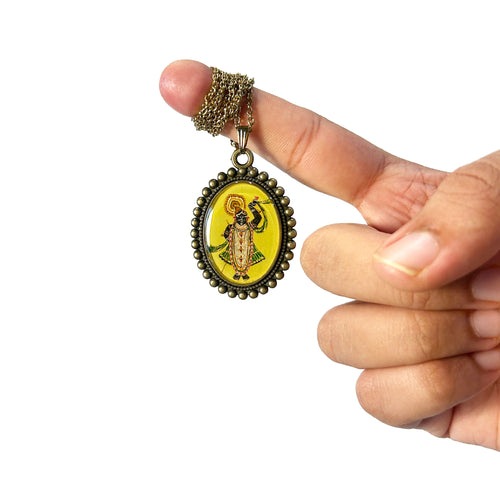 Hand holding a Shrinathji painting pendant with a lemon yellow background and intricate design.