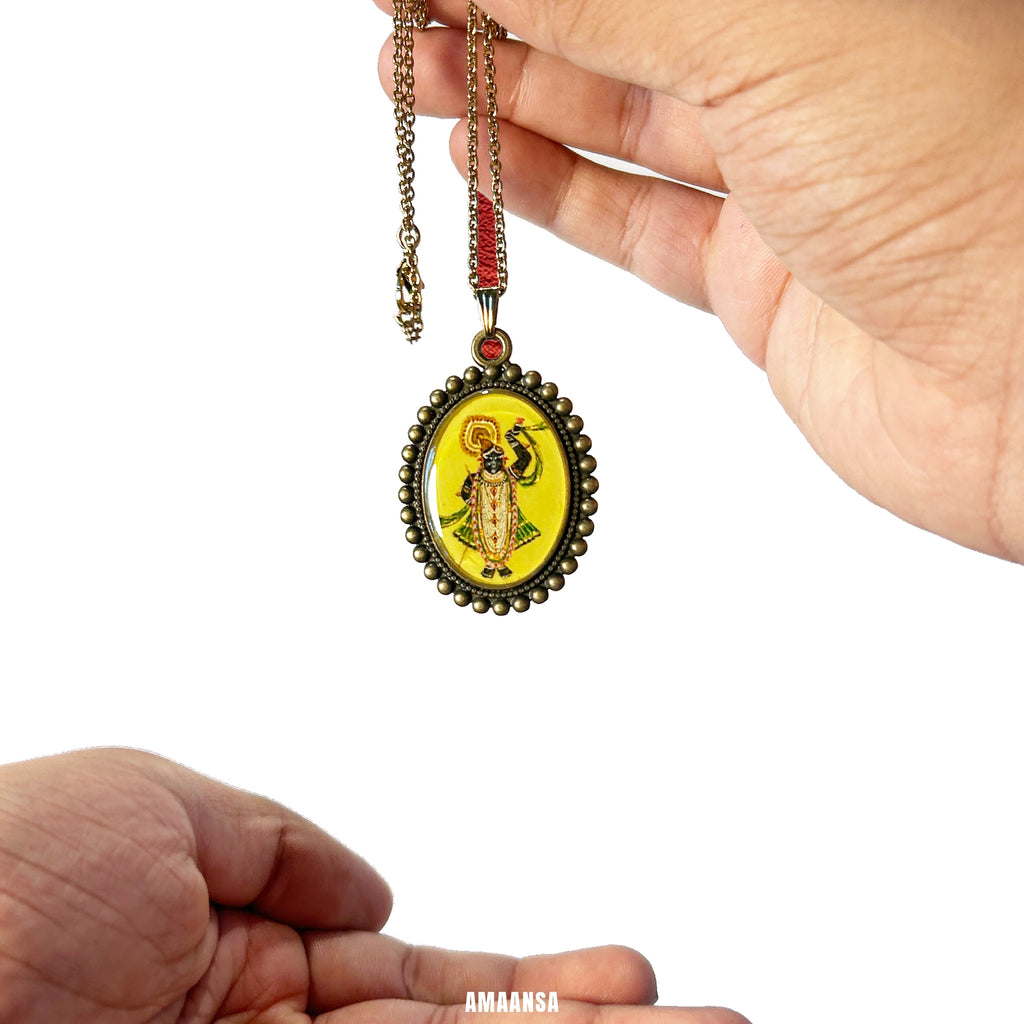 Hand holding a Shrinathji oval pendant on chain styled with soft fabric backdrop devotional Krishna gift necklace against a white background