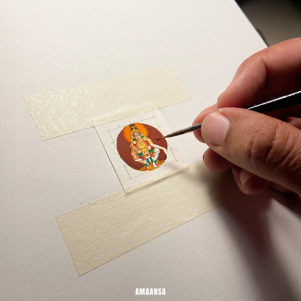 Hand holding a small brush over a miniature painting of Lord Ayyappa taped to board, brush adding details for devotional pendant on a white surface, with 'Amaansa' branding.