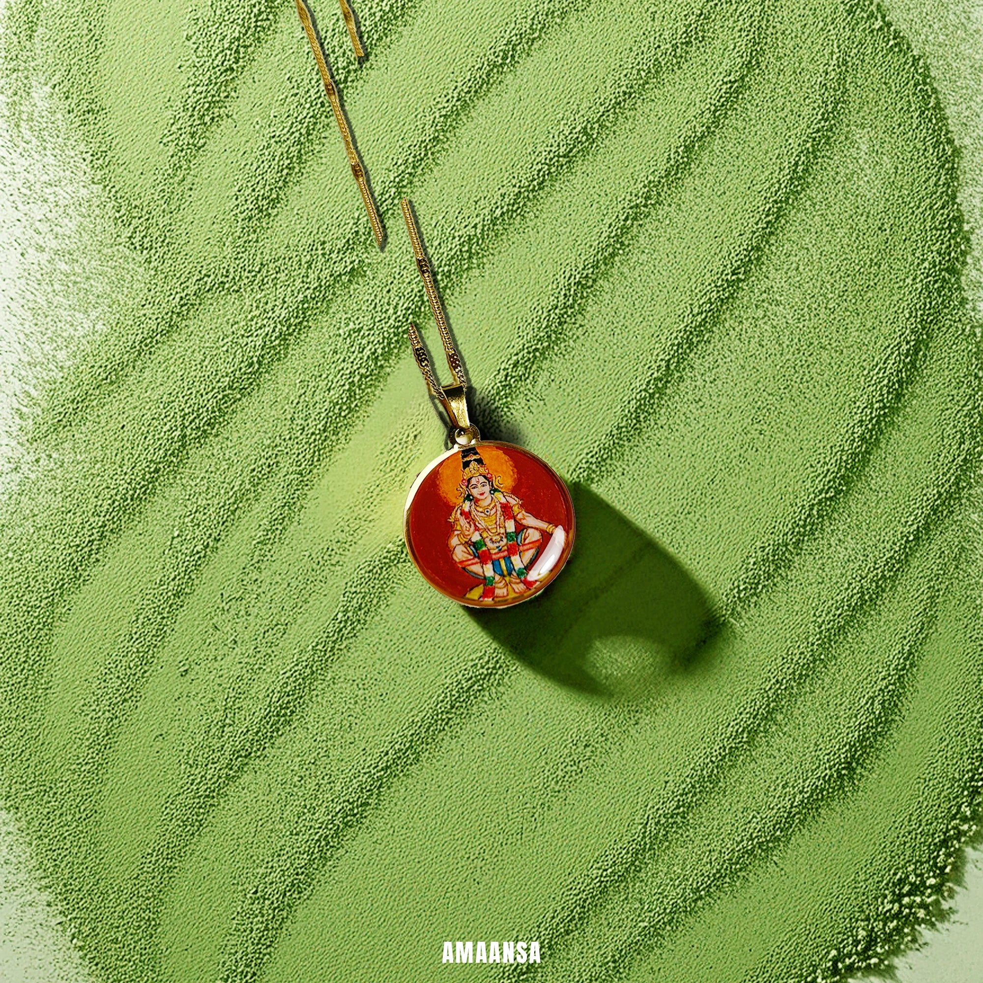a Round Lord Ayyappa pendant on a green textured surface, spiritual jewelry with 18k gold‑tone base.