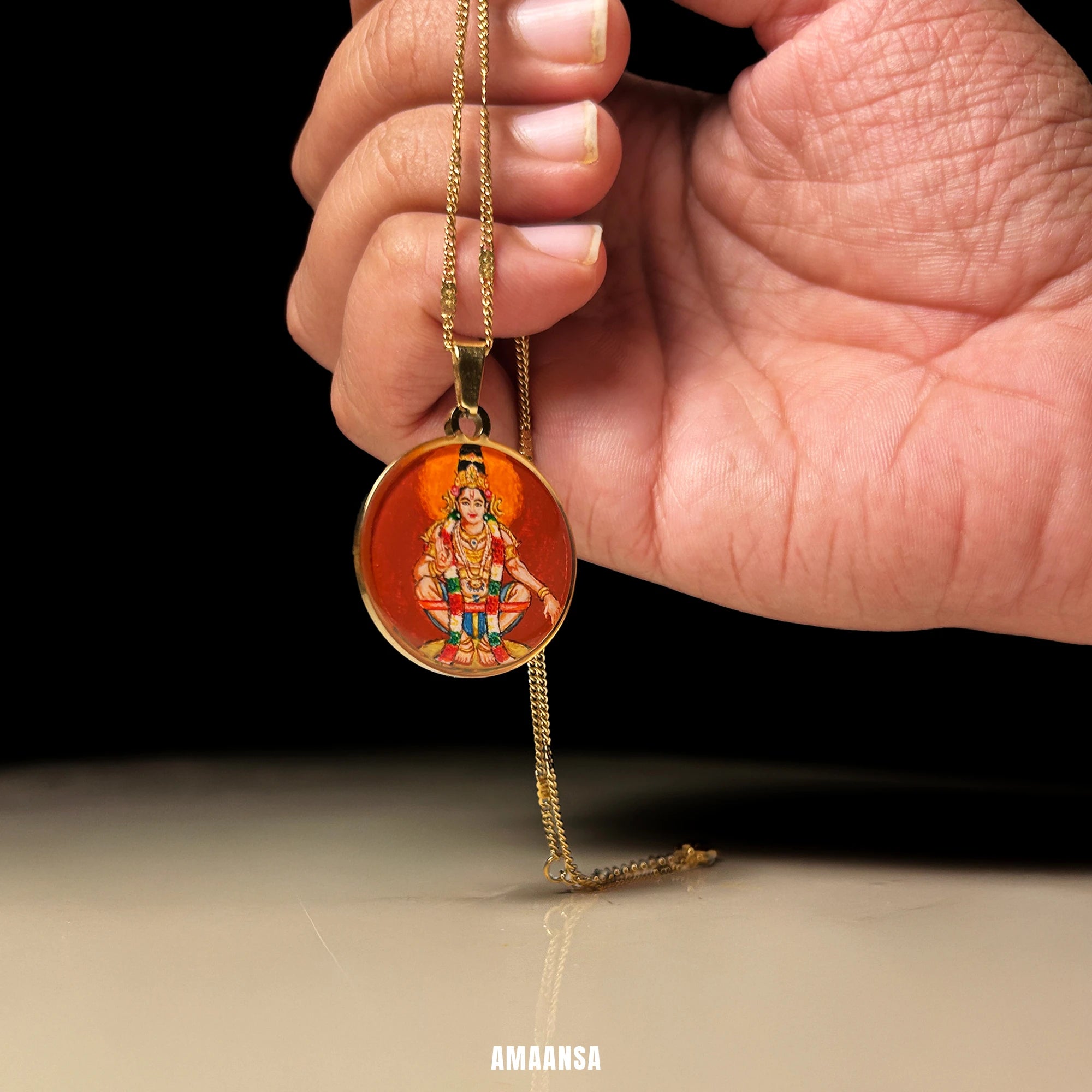 Hand holding a gold necklace with a pendant featuring Lord Ayyappa miniature painting pendant, on a neutral dark background.