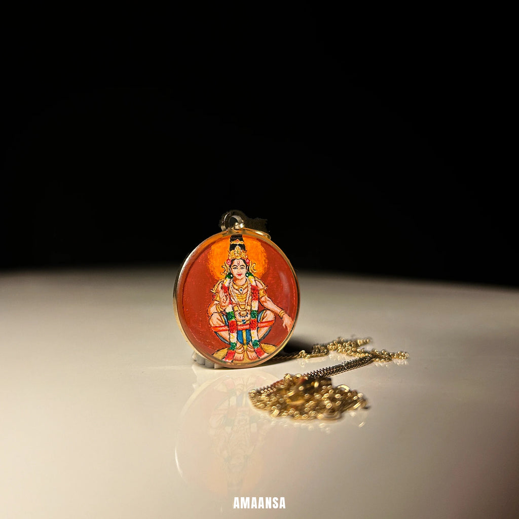 Close‑up of Lord Ayyappa pendant showing crown and ornaments, devotional necklace detail shot. Decorative painting pendant on an dark red-maroon background, displayed on a reflective surface.