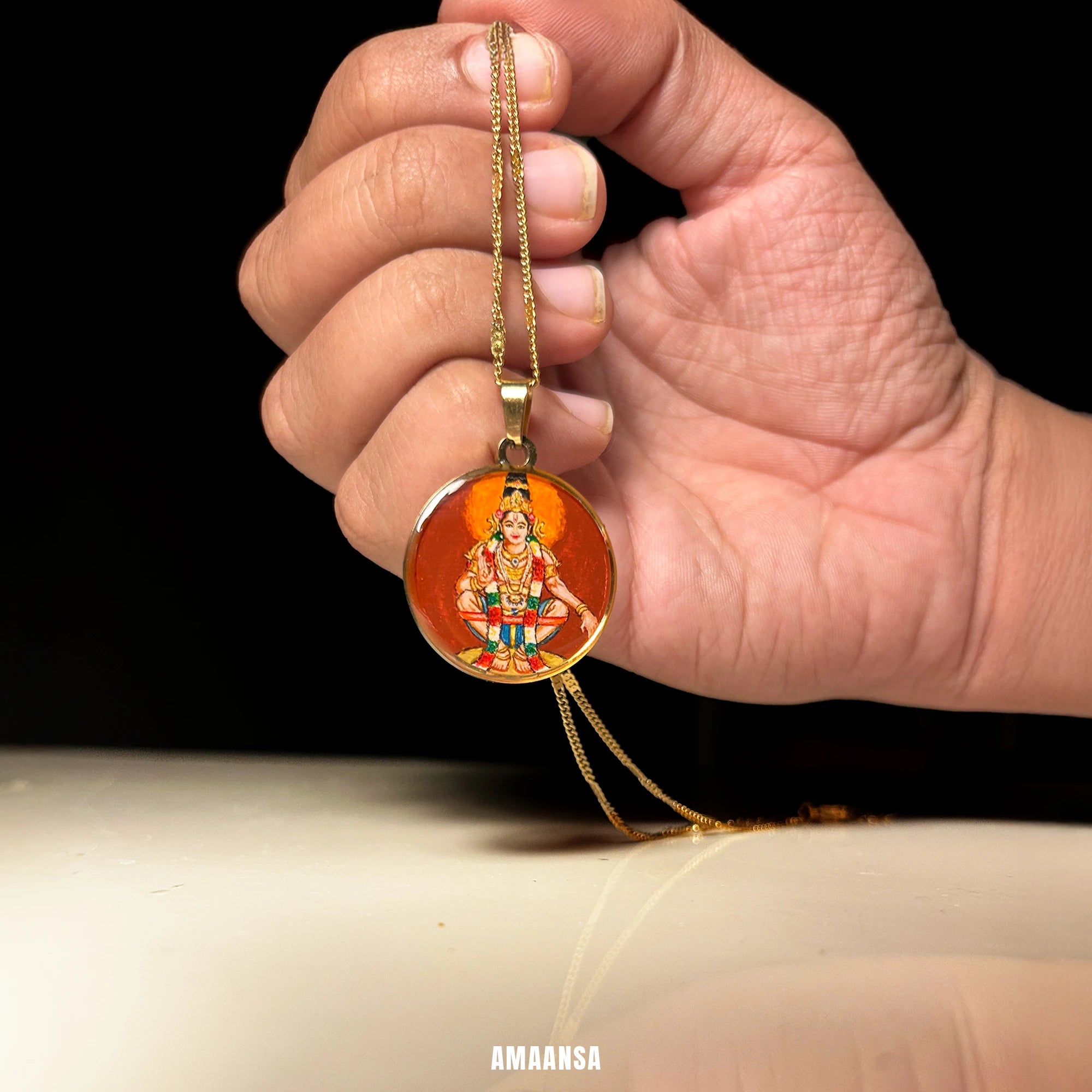 Hand holding a gold necklace with a miniature handpainted painting of Lord Ayyappa on chain, featuring an engraved design against a black background