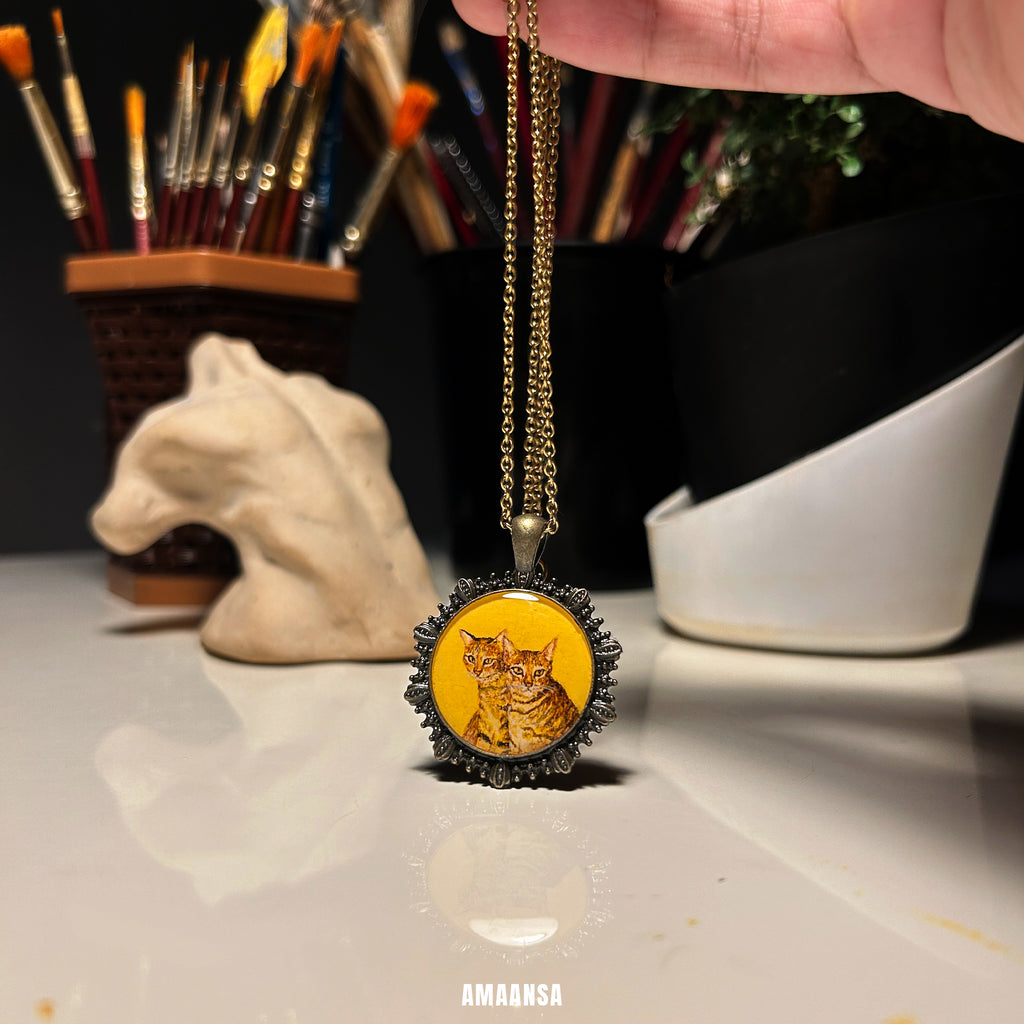 Necklace with a pendant featuring a 2 cats painting on a reflective surface with art supplies in the background. Custom two-face pet memorial pendant handpainted tabby and tortoiseshell cat miniature art vintage-style locket antique patina chain feline duo keepsake personalized tribute beloved companions collectible