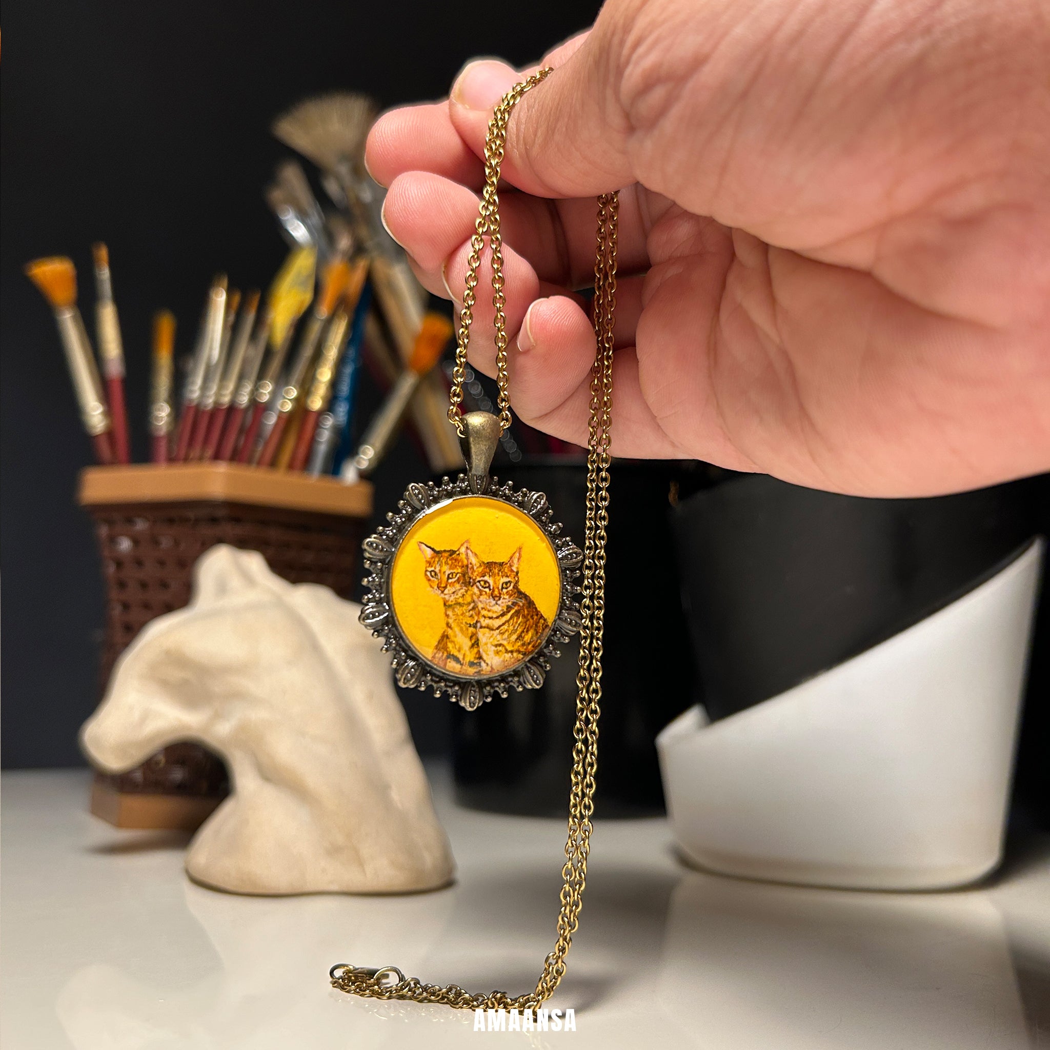 Hand holding a vintage necklace with a cat pendant in front of art supplies. Custom vintage locket handpainted domestic shorthair cat portrait antique patina chain feline companion keepsake personalized pet jewelry vintage yellow backdrop collectible remembrance tribute
