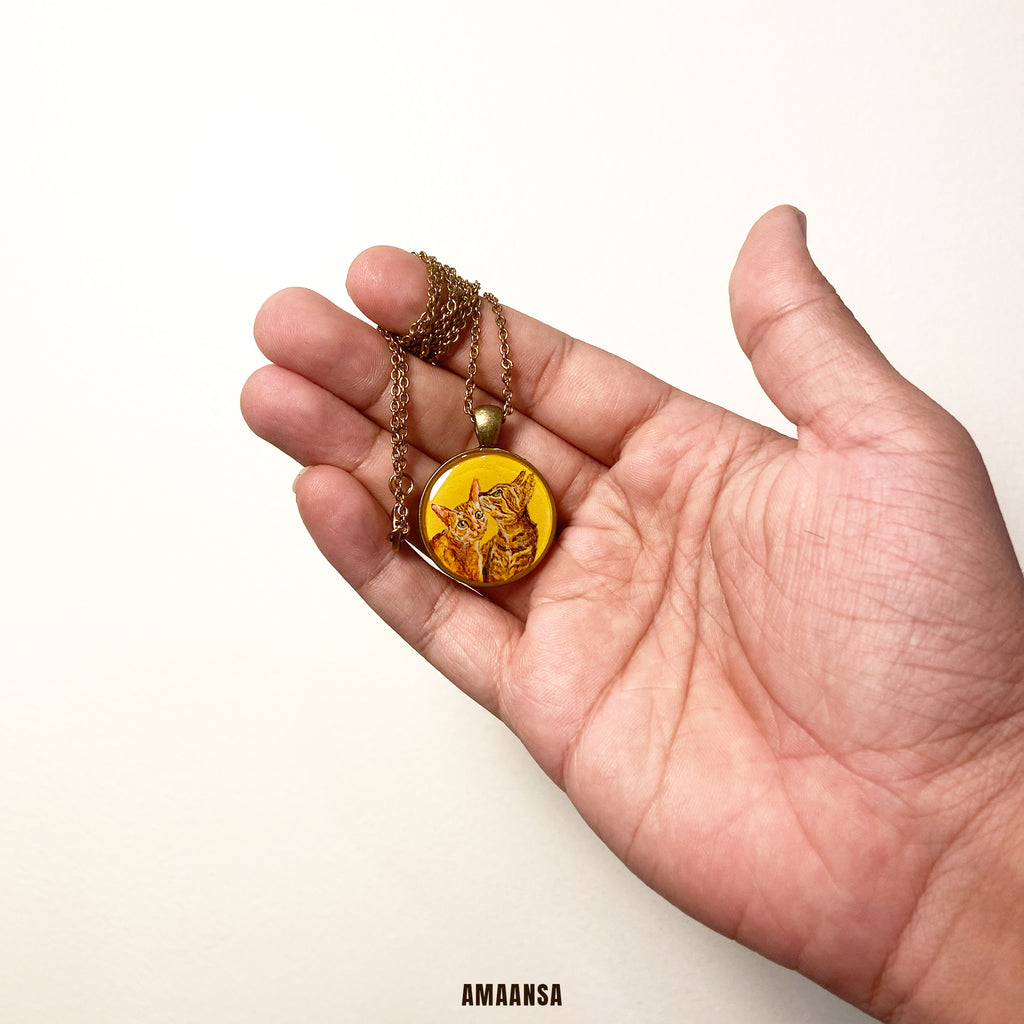 Handpainted two-face feline friends pendant vintage-style ornate chain cat duo miniature art personalized memorial locket custom pet jewelry antique finish keepsake collectible. Hand holding a necklace with a yellow pendant against a white background