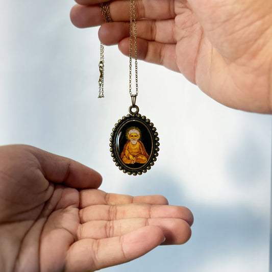 This miniature pendant features Guru Nanak ji in a meditative pose, with a radiant halo behind his head. Set against a black background, the oval-shaped painting captures his serene and spiritual expression. The pendant is encased in a vintage-style frame with a beaded outer rim, measuring 26mm x 32mm. A gold-tone chain complements the piece, offering a wearable art form from AMAANSA Fine Arts Studio.