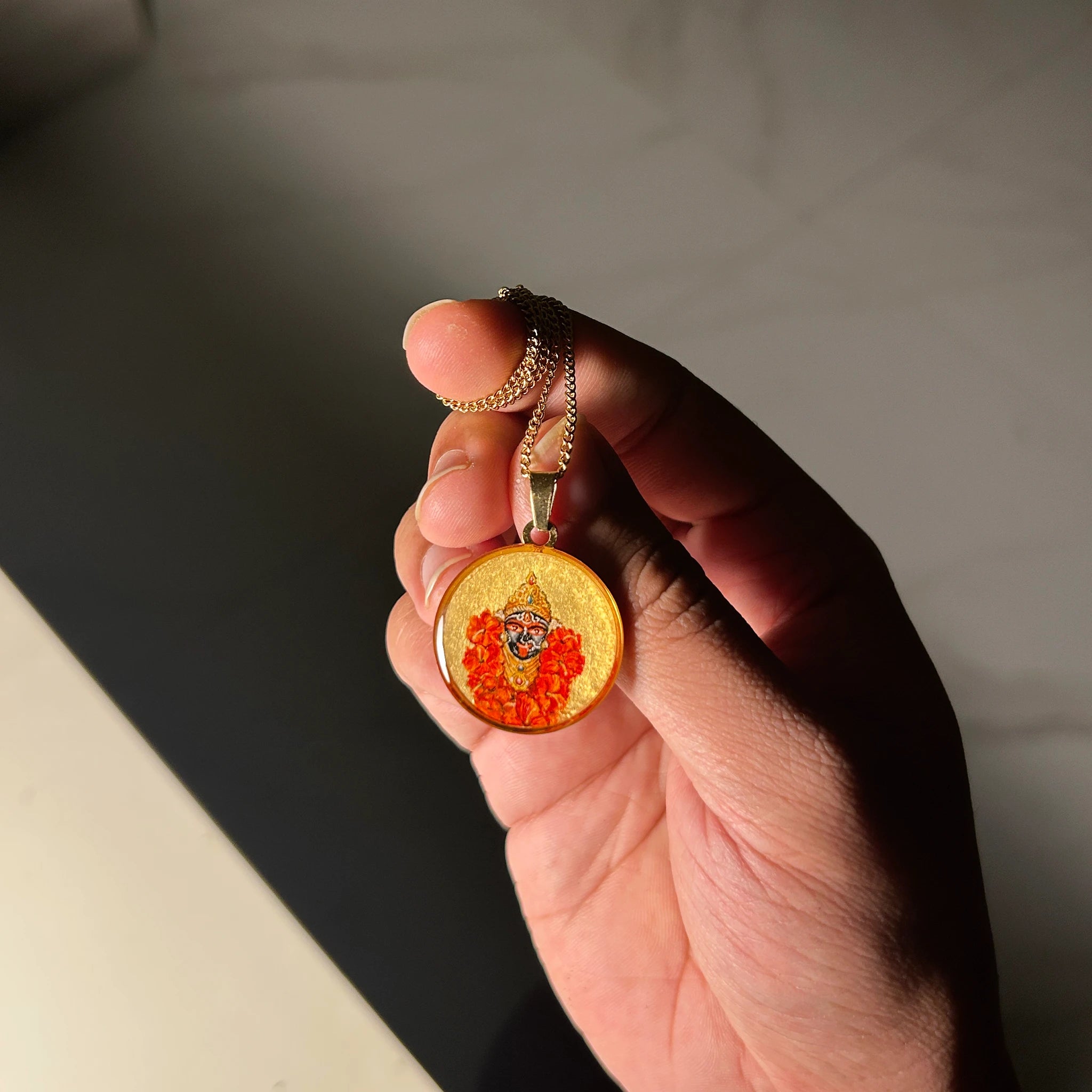 Maa Kali 18k gold‑plated miniature portrait pendant on chain, gold‑tone frame resting on sunlit hand surface for spiritual jewelry display.
