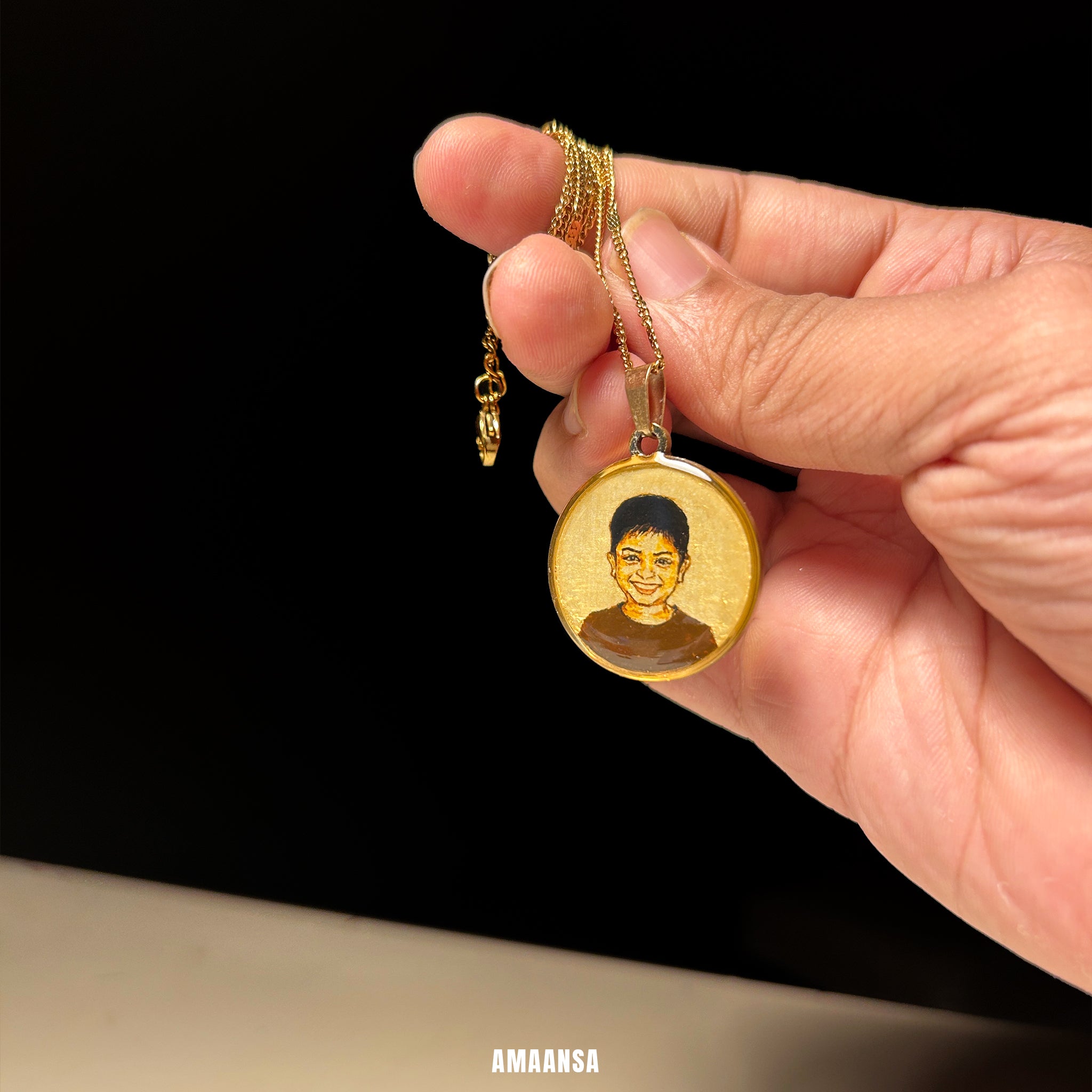 Hand holding a gold necklace with a pendant featuring a portrait, against a black background. Custom 18k gold PVD portrait pendant four year old toddler handpainted miniature art mother's love luxury jewelry stainless steel base burnt umber hues heirloom keepsake parental treasure AMAANSA artist