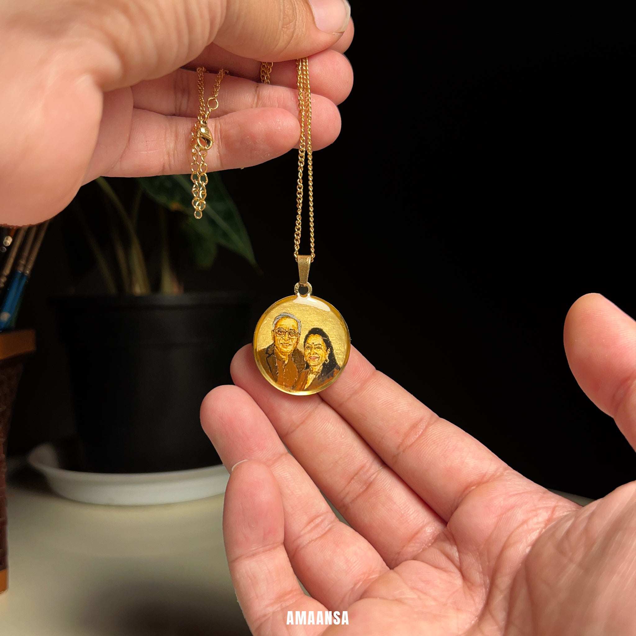 Gold pendant with a family portrait held by a hand against a black background. Custom 18k gold portrait pendant affluent middle age couple handpainted miniature art luxury heirloom jewelry wife commissioned burnt umber painting stainless steel premium craftsmanship AMAANSA artistry treasure