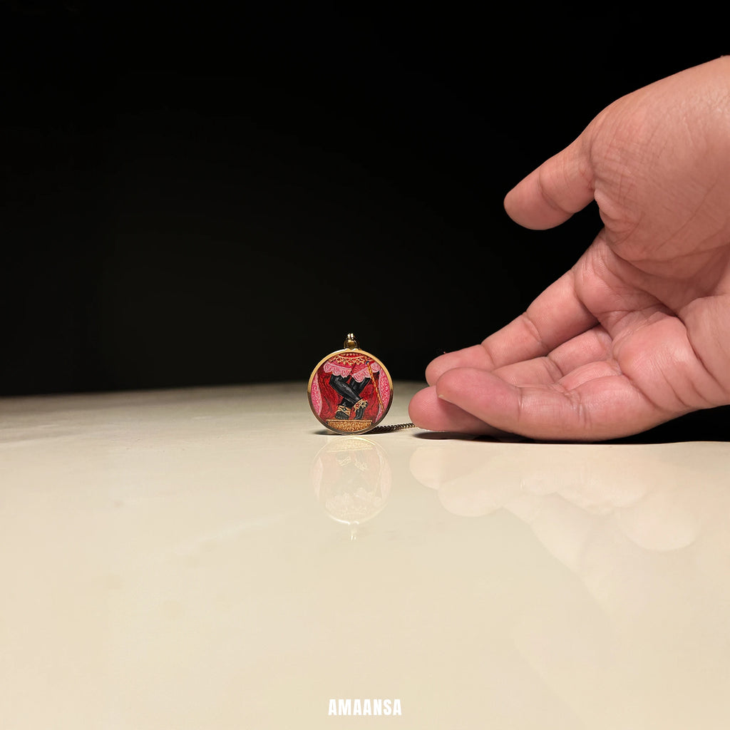 18k gold‑plated Sri Radharamanji paduka pendant on a reflective surface with a hand reaching towards it, branded 'AMAANSA'. Hand shows size comparison