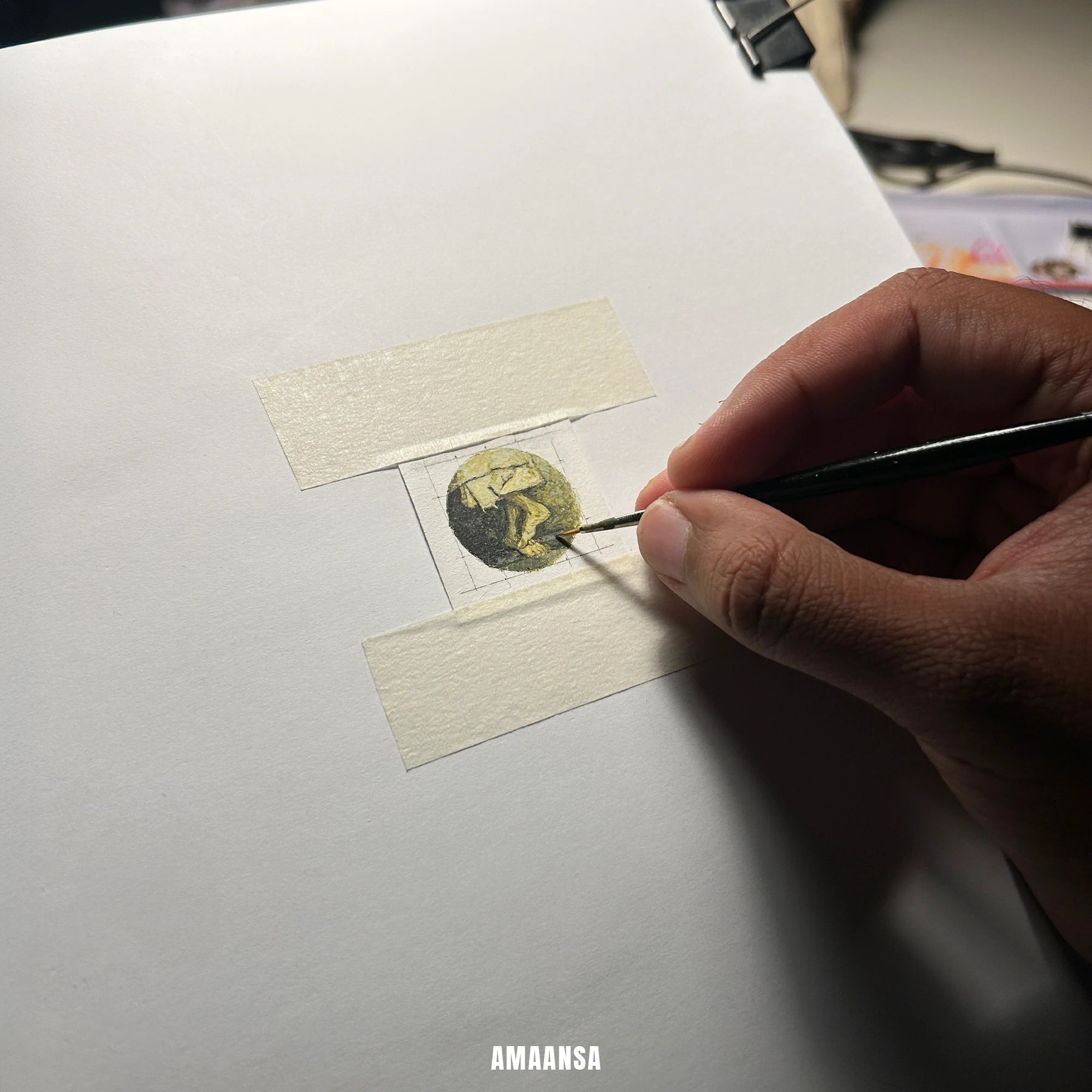 Arist using a brush to work on a small painting or artwork on a white surface. Finished Sai Baba charan miniature painting on taped square, circular sacred feet artwork ready to be set into 18k gold‑plated pendant.