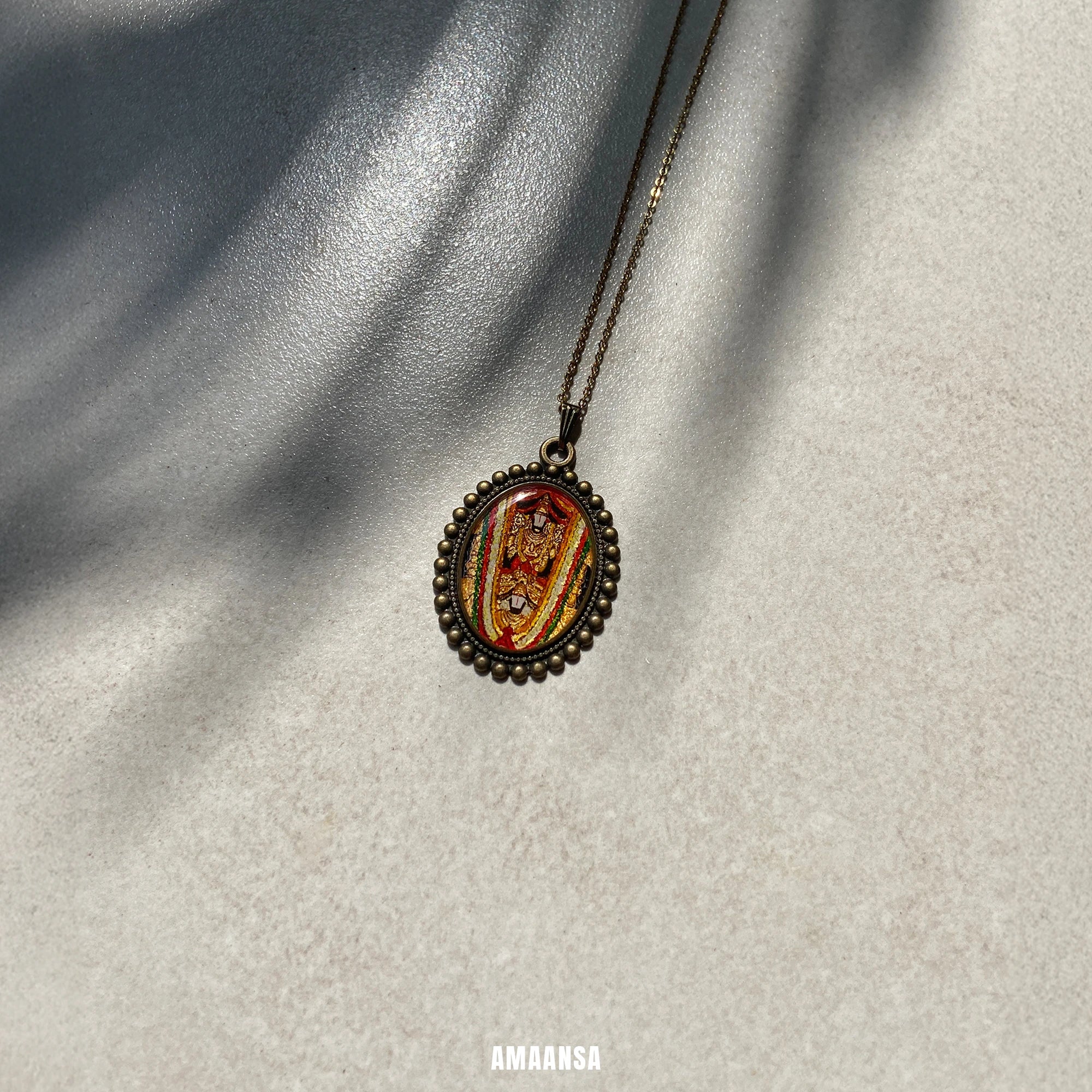 Venkateswara Swamy antique gold‑tone oval miniature pendant on chain, hand‑painted devotional jewelry photographed on sunlit stone background on a textured surface, brand 'AMAANSA' visible