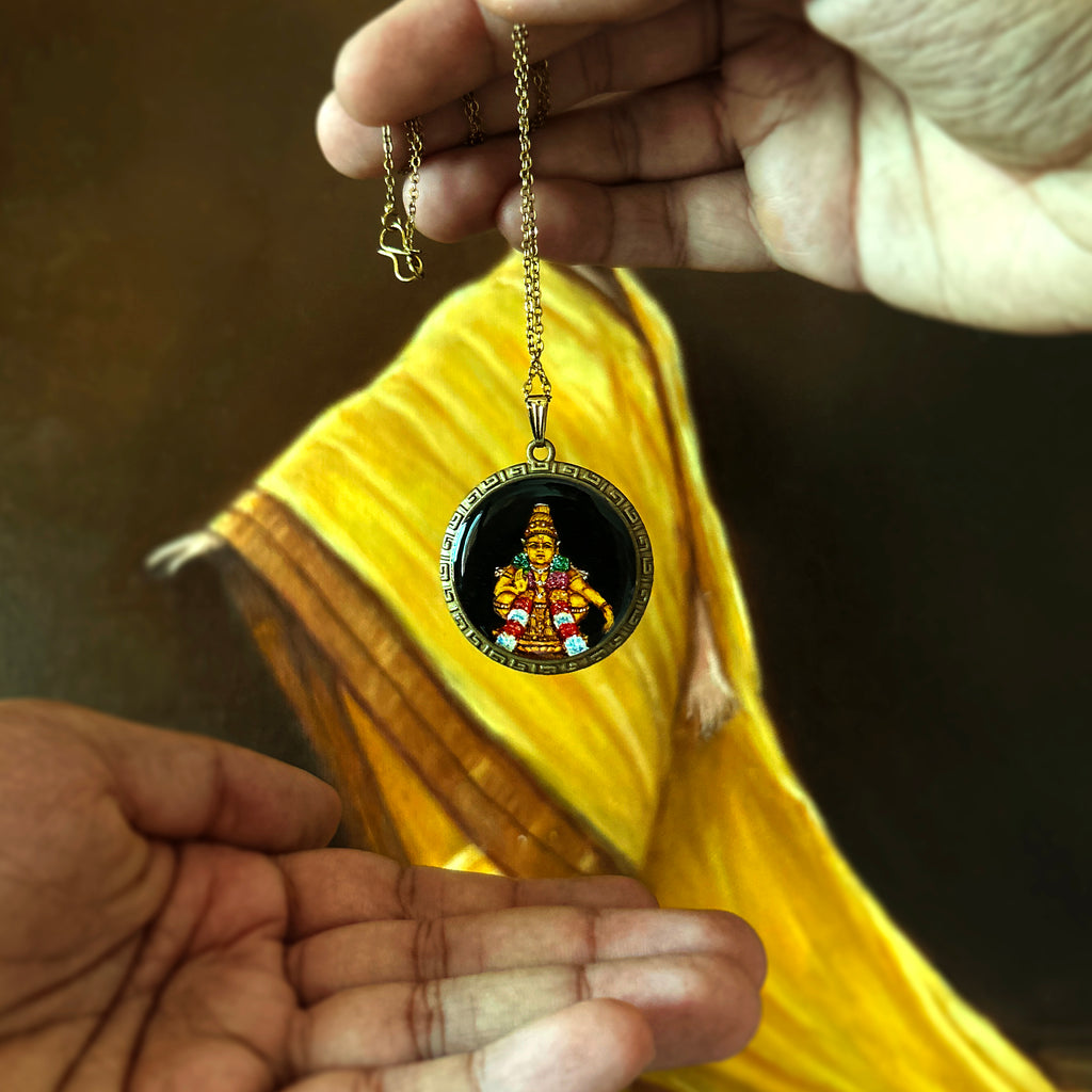 Artistry in Faith Collection - 30mm Lord Ayyappa pendant suspended between two hands against vibrant yellow fabric oil-painting background, displaying delicate antique brass chain with S-hook clasp. Hand-painted deity with intricate ceremonial details illuminated on mystical black background, framed by vintage Greek key pattern. Two hands frame the pendant's gentle sway, creating dramatic composition that highlights both devotional significance and artisan craftsmanship