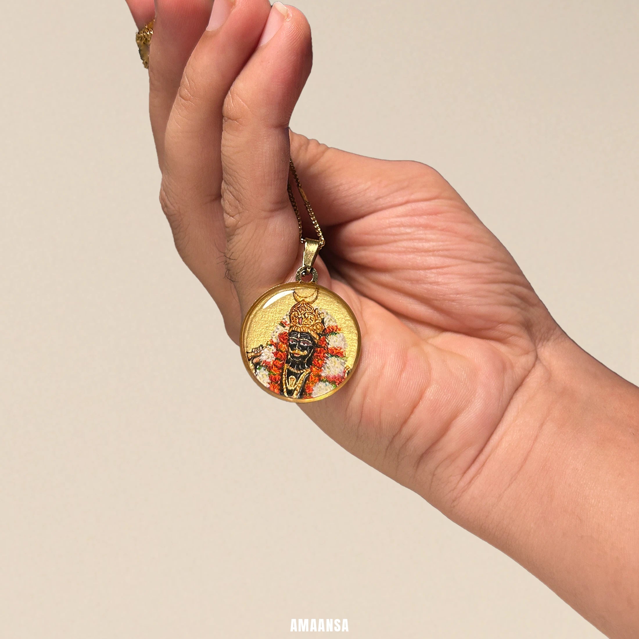 Hand holding a Kaal Bhairava 18k gold‑plated miniature portrait pendant on chain, devotional necklace with intricate hanging agains a beige background