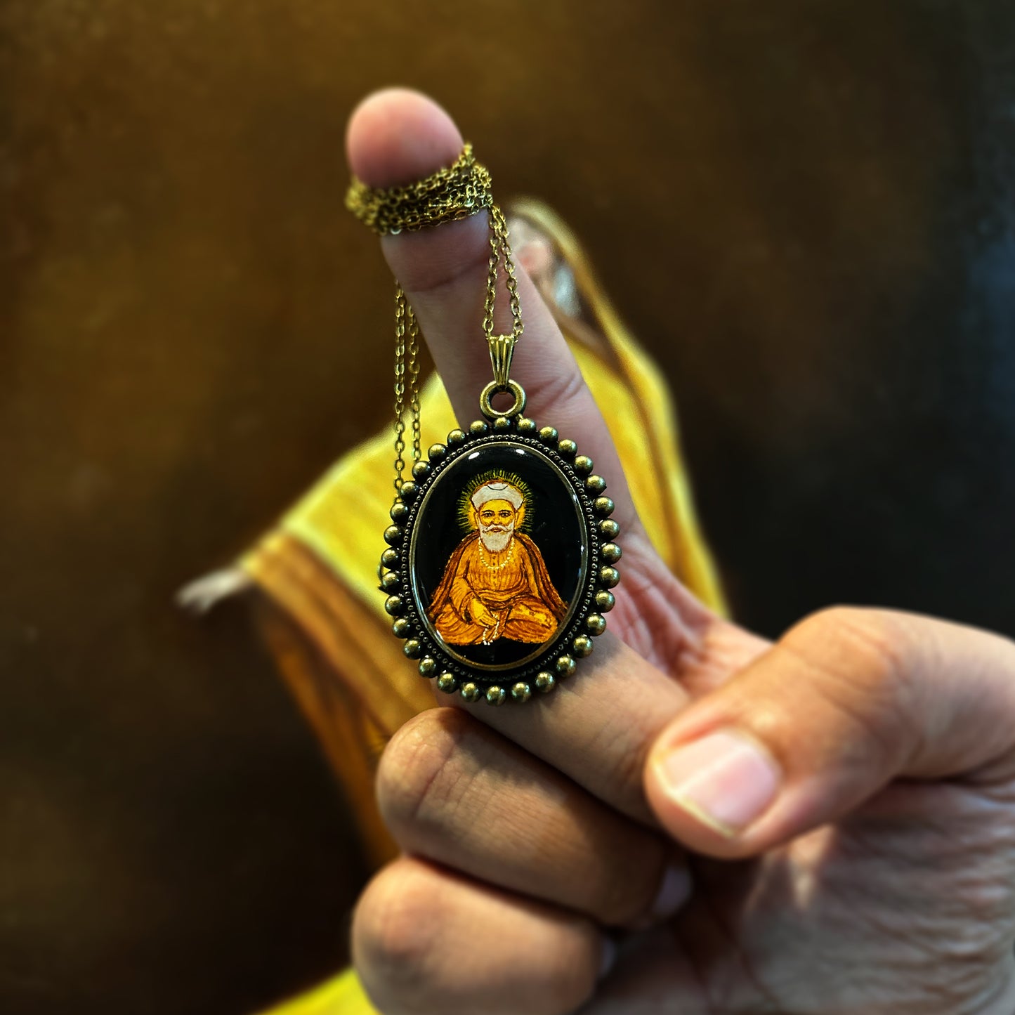 Miniature painting of Guru Nanak ji in an upright meditative posture, with a halo radiating from behind his head. The black background emphasizes the calm, spiritual energy of the piece. This oval-shaped pendant, measuring 26mm x 32mm, features a vintage-style frame with a delicate bead design. Paired with a chain, this piece from AMAANSA Fine Arts Studio is a tribute to divine meditation and tranquility.