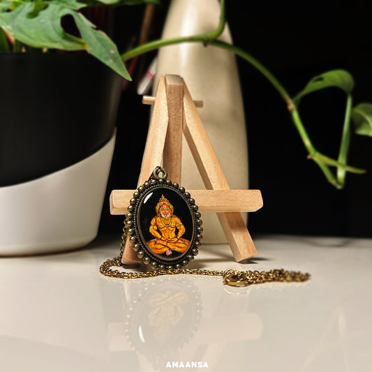 This wearable art piece from AMAANSA Fine Arts Studio's 'Artistry in Faith' collection depicts Lord Hanuman seated in meditation. Painted on a 26mm x 32mm oval base with a black background, the portrait uses vintage yellow hues to highlight his crown, meditative hands, and a small flower at his feet. The pendant features a moti-style metal design along the rim, enhancing its vintage aesthetic. Accompanied by a chain, this miniature painting is both spiritual and decorative. 