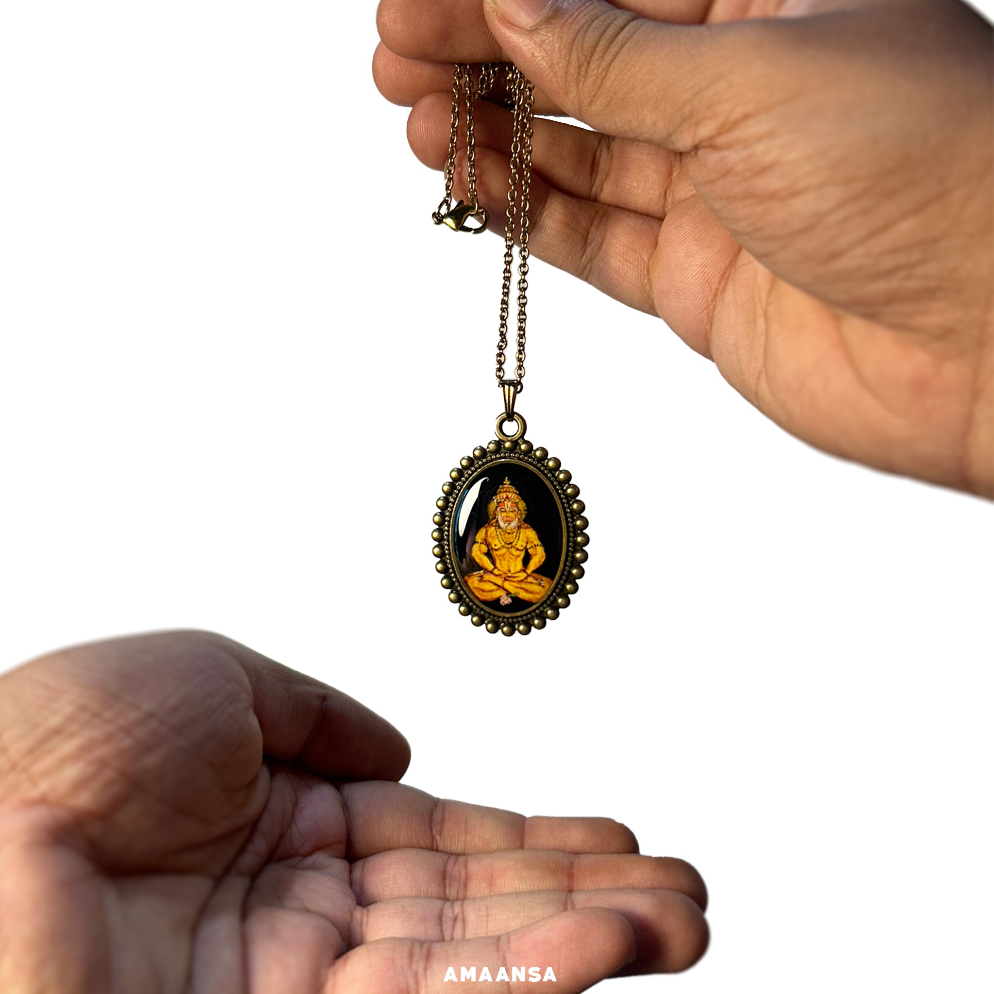 An oval-shaped pendant showcasing Lord Hanuman from the 'Artistry in Faith' collection, created as a 26mm x 32mm miniature painting in vintage yellow hues. Hanumanji, wearing a crown and with a divine glow behind him, sits in meditation, hands resting near his legs with a small flower below. The black background enhances the painting's contrast, while the outer rim features a vintage moti design. Paired with a chain as a wearable art. Painting is protected by a metal base and resin coating.