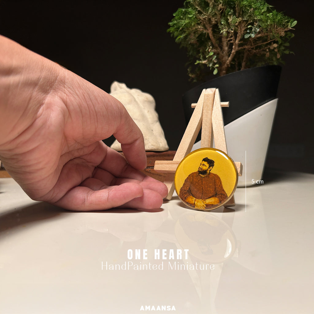 AMAANSA Fine Arts Studio's compact 5cm heirloom portrait by artist Abhinav - capturing young man's genuine smile and rugged beard in casual attire, a heartfelt engagement gift handpainted in warm sepia and burnt umber against vintage yellow, artist's hand demonstrating the miniature scale beside wooden easel and bonsai backdrop