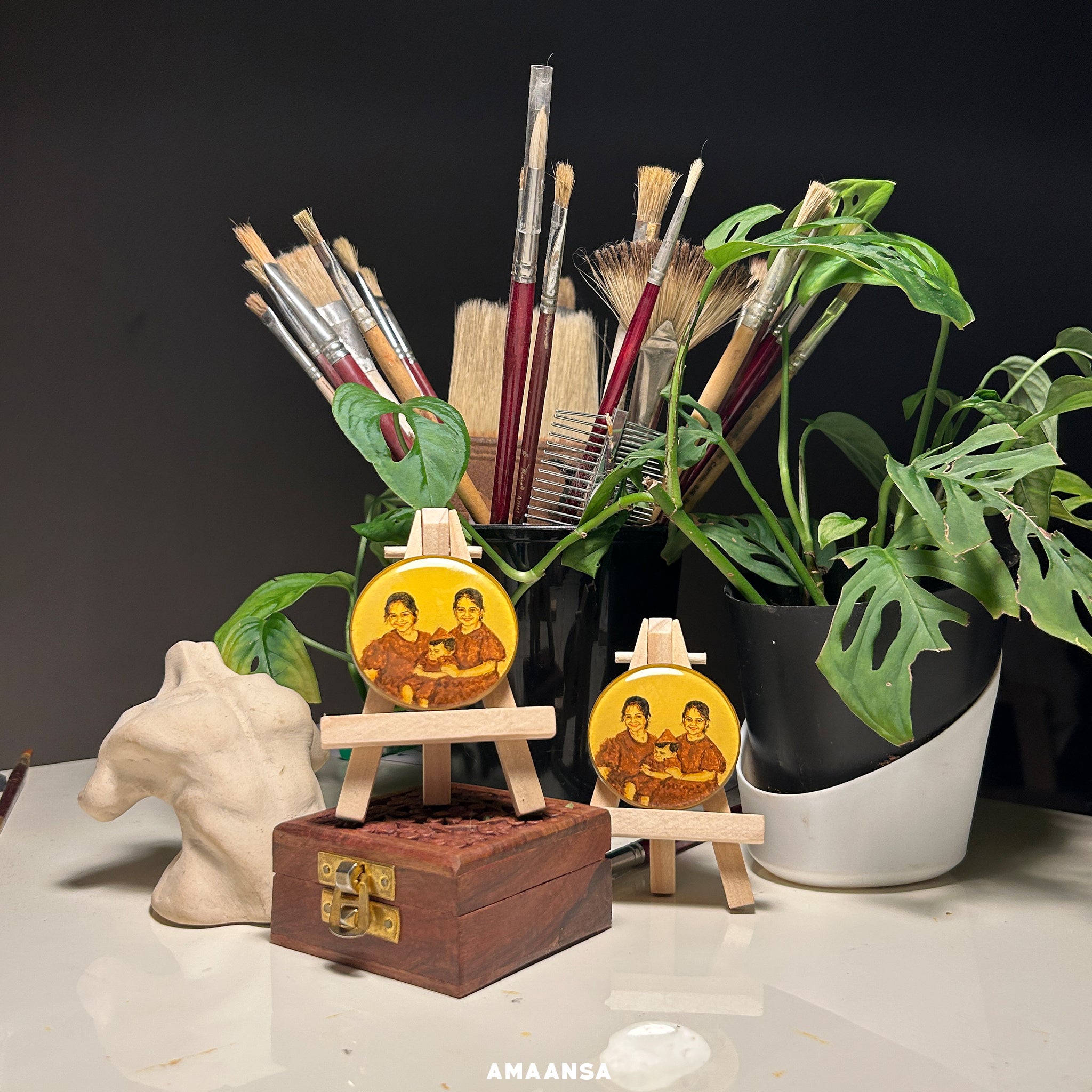 Twin sisters lovingly embracing their baby brother, reimagined by AMAANSA Fine Arts Studio artist Abhinav in 50mm twin heirloom pieces - identical bright smiles and matching brown outfits transformed from vintage black and white photograph, each miniature delicately handpainted in rich sepia tones against luminous vintage yellow, artistically staged with antique paintbrushes, carved trinket box, and lush monstera leaves framing the scene