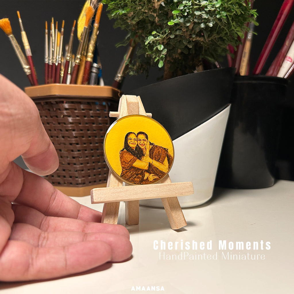AMAANSA Fine Arts Studio presents 50mm handpainted heirloom by artist Abhinav, capturing tender mother-daughter duo with matching smiles and brown attire, daughter's flowing hair framing her face as she embraces her mother, meticulously rendered in sepia and burnt umber against vintage yellow backdrop, displayed in artist's studio amid paintbrushes and bonsai