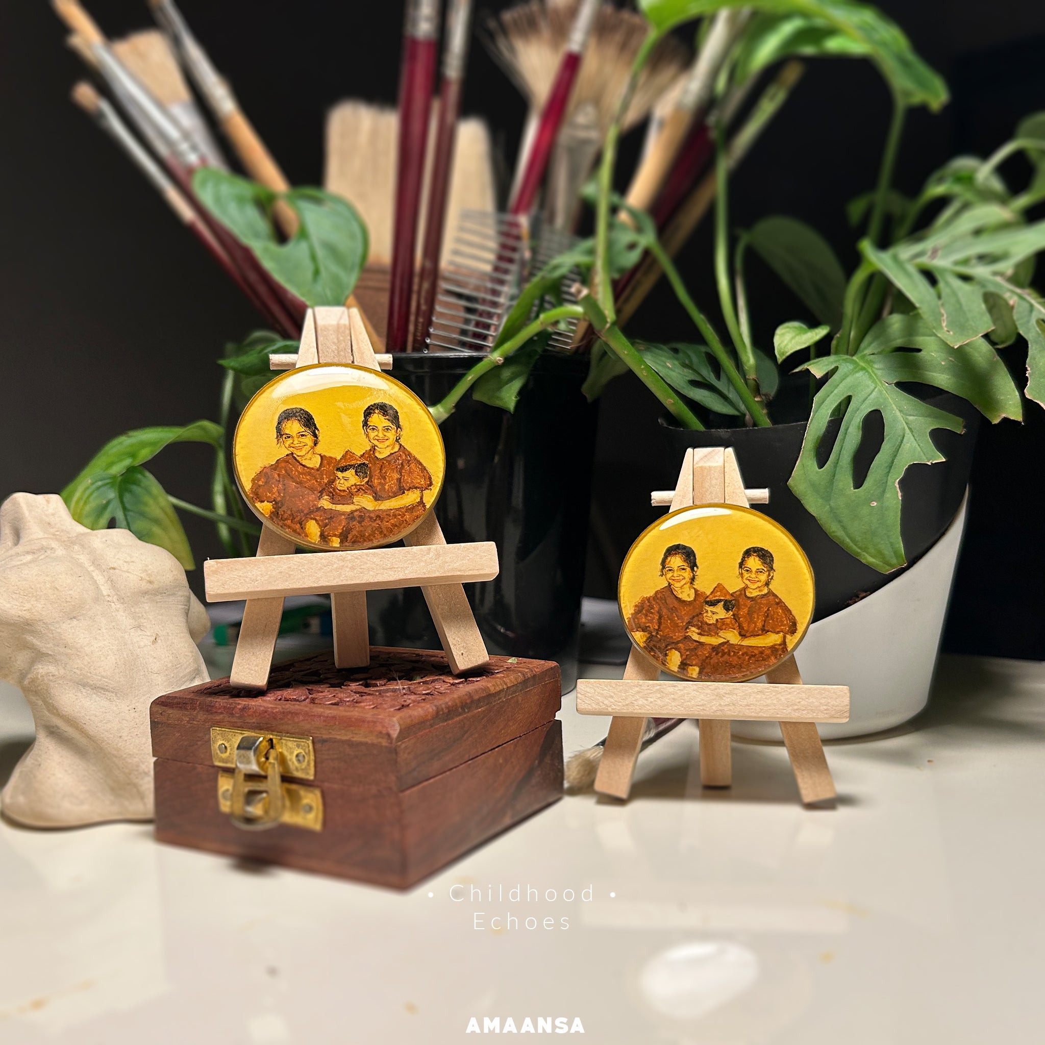 Nostalgic 50mm heirloom portraits by AMAANSA Fine Arts Studio artist Abhinav, recreating cherished black and white photograph of twin sisters with matching smiles cradling their little brother, expertly rendered in sepia and burnt umber against vintage yellow, dual pieces displayed on wooden easels amid artist's paintbrushes, carved wooden box, and vibrant monstera leaves