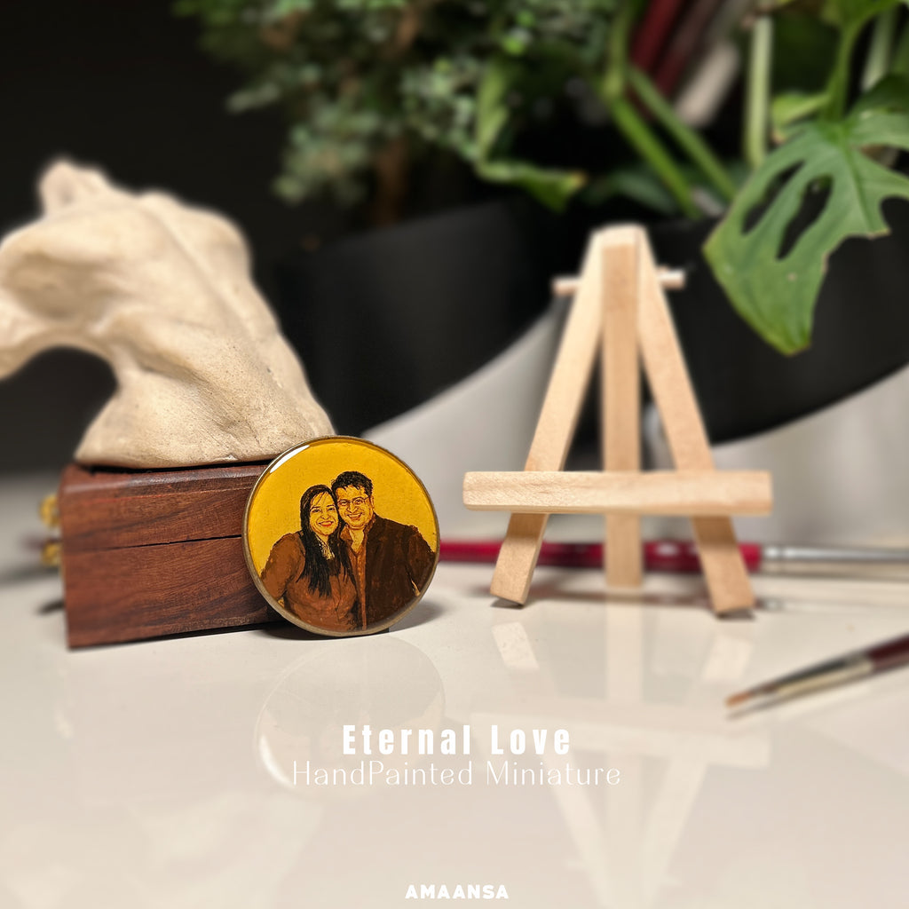 Handcrafted 50mm heirloom portrait from AMAANSA Fine Arts Studio capturing young couple's joyful bond - her serene smile and long dark hair complementing his warm expression and glasses, their faces close in loving embrace, meticulously painted by artist Abhinav in sepia and burnt umber against vintage yellow, staged with artist's tools - paintbrushes and wooden easel beside decorative ceramic piece and monstera leaves