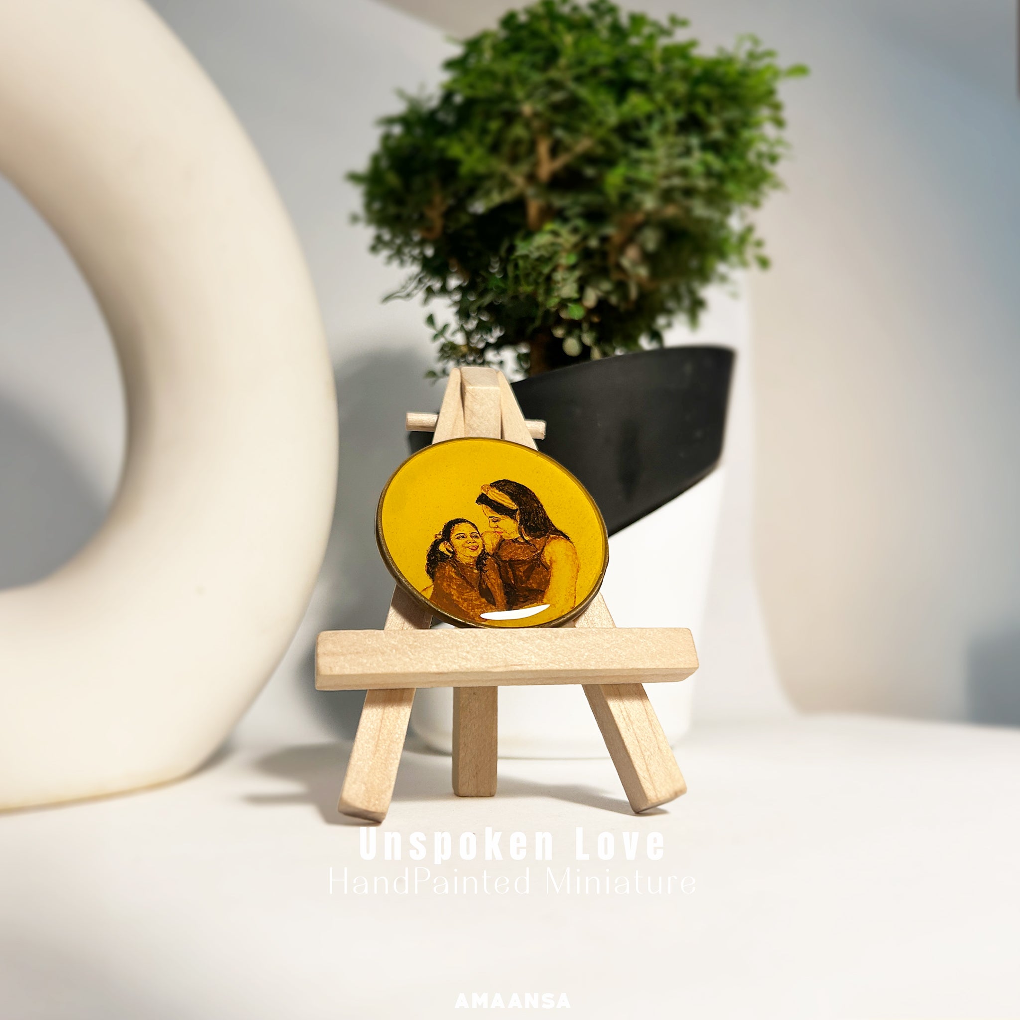 AMAANSA Fine Arts Studio presents an intimate 50mm heirloom portrait of mother and daughter sharing a tender moment, daughter's radiant smile as mother gazes down lovingly, their long flowing hair framing their faces, delicately handpainted by artist Abhinav in sepia and burnt umber tones against vintage yellow background, displayed on wooden easel with minimalist ceramic and bonsai decor