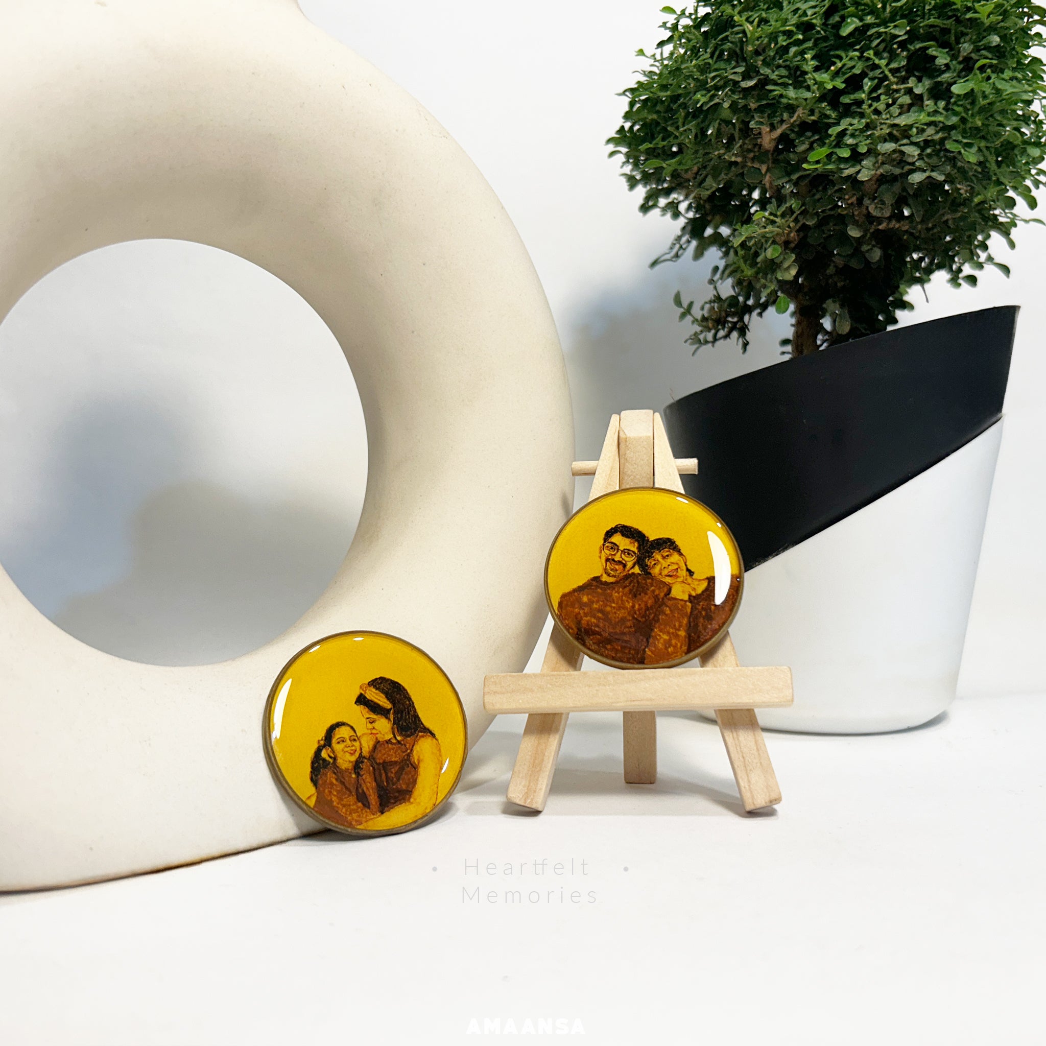 Handcrafted by artist Abhinav at AMAANSA Fine Arts Studio, dual 50mm miniature portraits showcasing intimate moments - a young couple sharing a warm embrace with his glasses and gentle smile, alongside a mother-daughter portrait with flowing hair and shared laughter, both painted in sepia and burnt umber against vintage yellow, artistically staged with wooden easel and minimalist decor