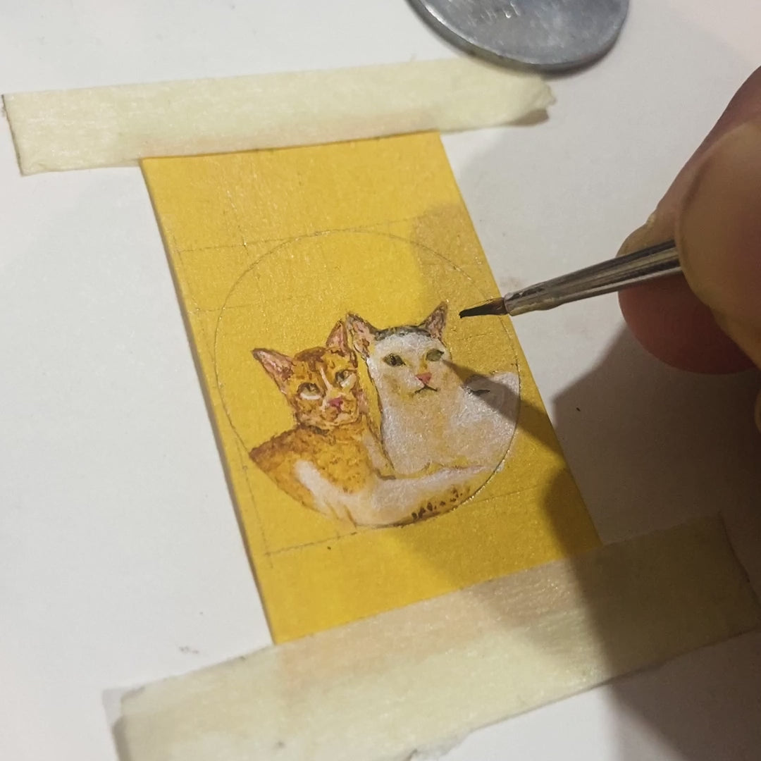 25-second craftsman video revealing delicate miniature creation: artist's hand skillfully detailing two beloved cats - ginger tabby and white companion nestled together, each tiny face smaller than artist's fingernail. One-rupee coin provides scale reference as finest brush defines whiskers and expressions on naples yellow canvas, later transformed into pendant with artisan resin coating. Remarkable precision demonstrated where each feline's features, from alert ears to gentle eyes, occupy mere millimeters.