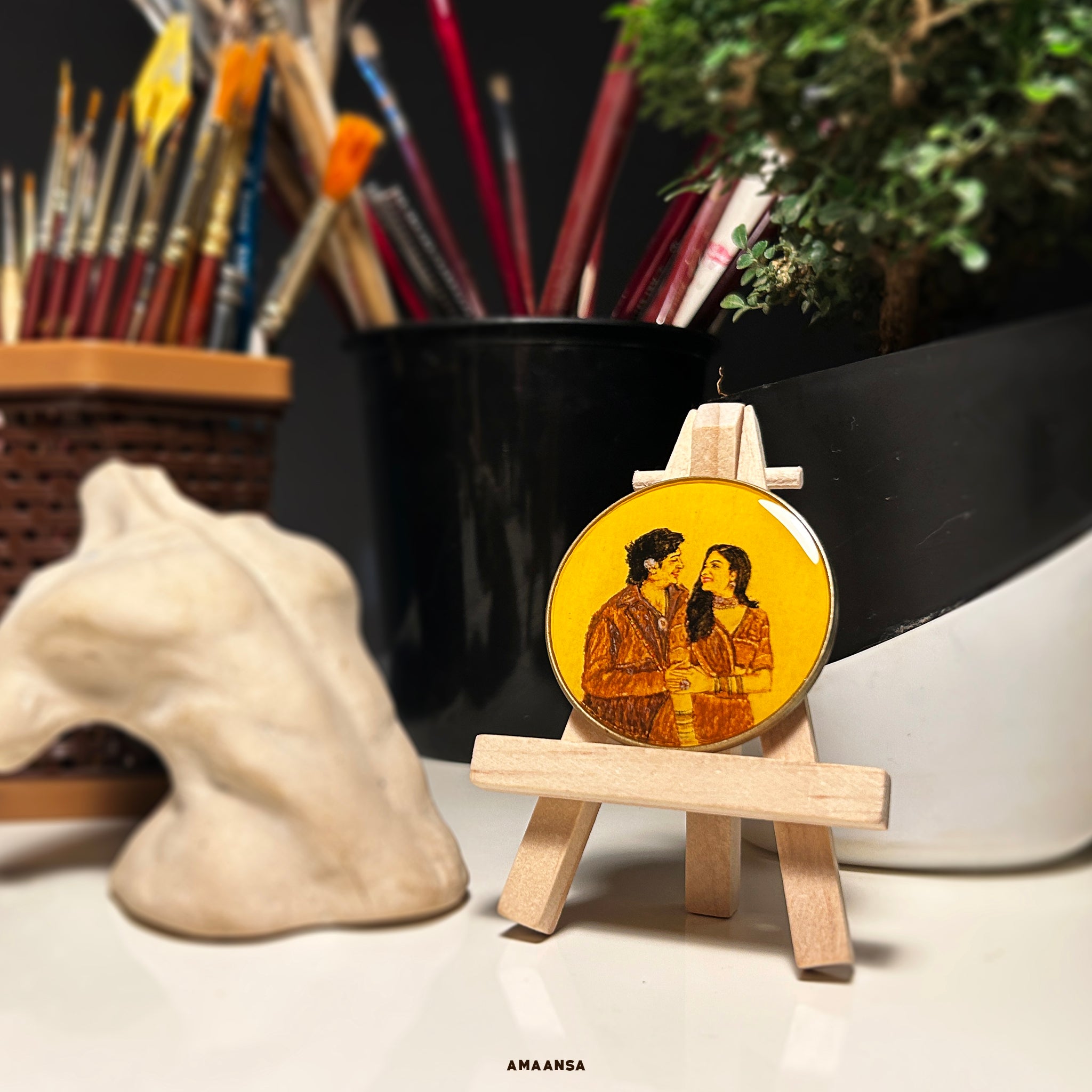 Smaran Collection - Handcrafted custom miniature portrait (5cm) capturing couple in traditional Gujarati Garba attire, meticulously painted with vintage tones on naples yellow background. Elegantly displayed on wooden easel against dramatic black backdrop with lush greenery and curated decor, creating an artistic tabletop vignette. Intimate moment between gazing couple preserved as cherished heirloom-quality art by AMAANSA Fine Arts Studio