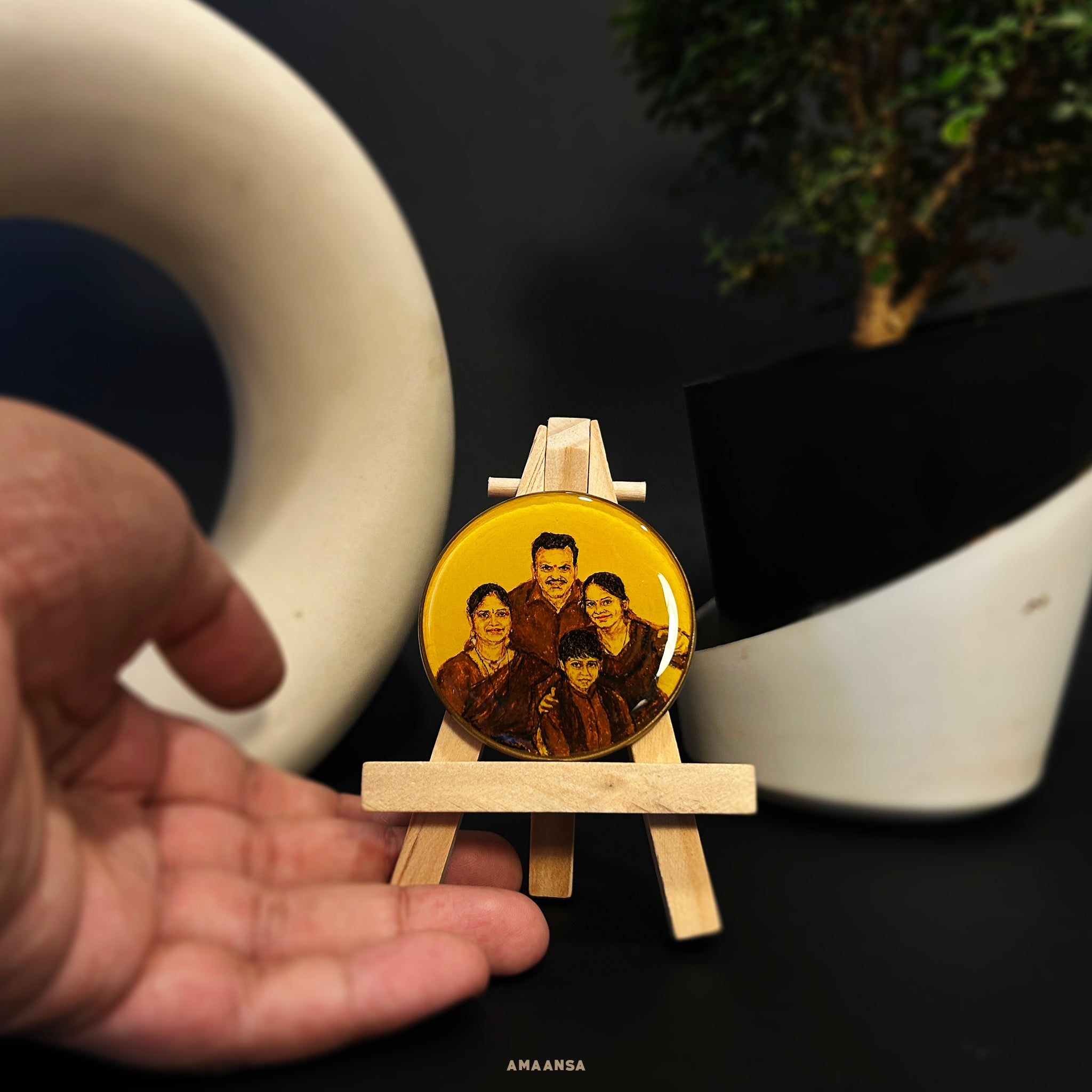 Handpainted family portrait miniature (5cm) capturing tender family moment: father standing protectively behind with gentle smile, mother seated with warm expression, daughter and son nestled close with bright faces, all painted in rich brown tones on naples yellow background. Artfully displayed on wooden easel amid contemporary black-white decor and lush greenery, transforming intimate family bonds into cherished tabletop heirloom by AMAANSA Fine Arts Studio, where each expression tells its own story of lo