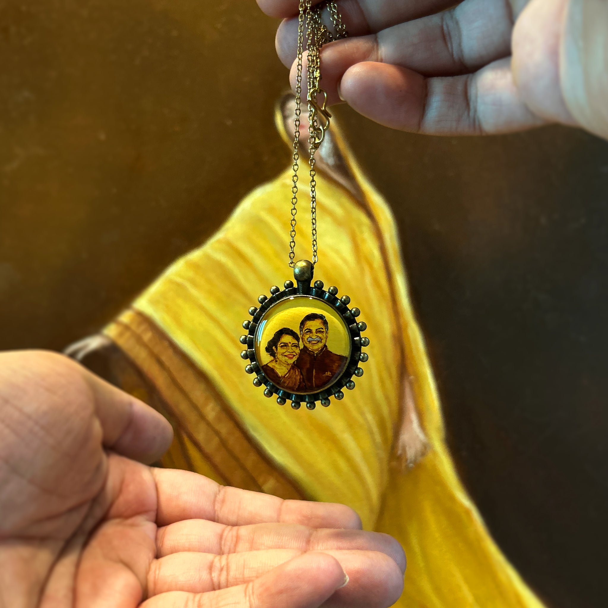 Meticulously crafted anniversary keepsake (26mm) celebrating 25 years: parents' joyful expressions captured in sepia tones, mom in traditional attire beside dad's warm smile. Pendant Portrait suspended between palms against artist's luminous oil painting, beaded border framing this milestone moment painted in naples yellow for timeless appeal. Each loving detail preserved beneath protective finish, antique gold colored chain completing this cherished gift.