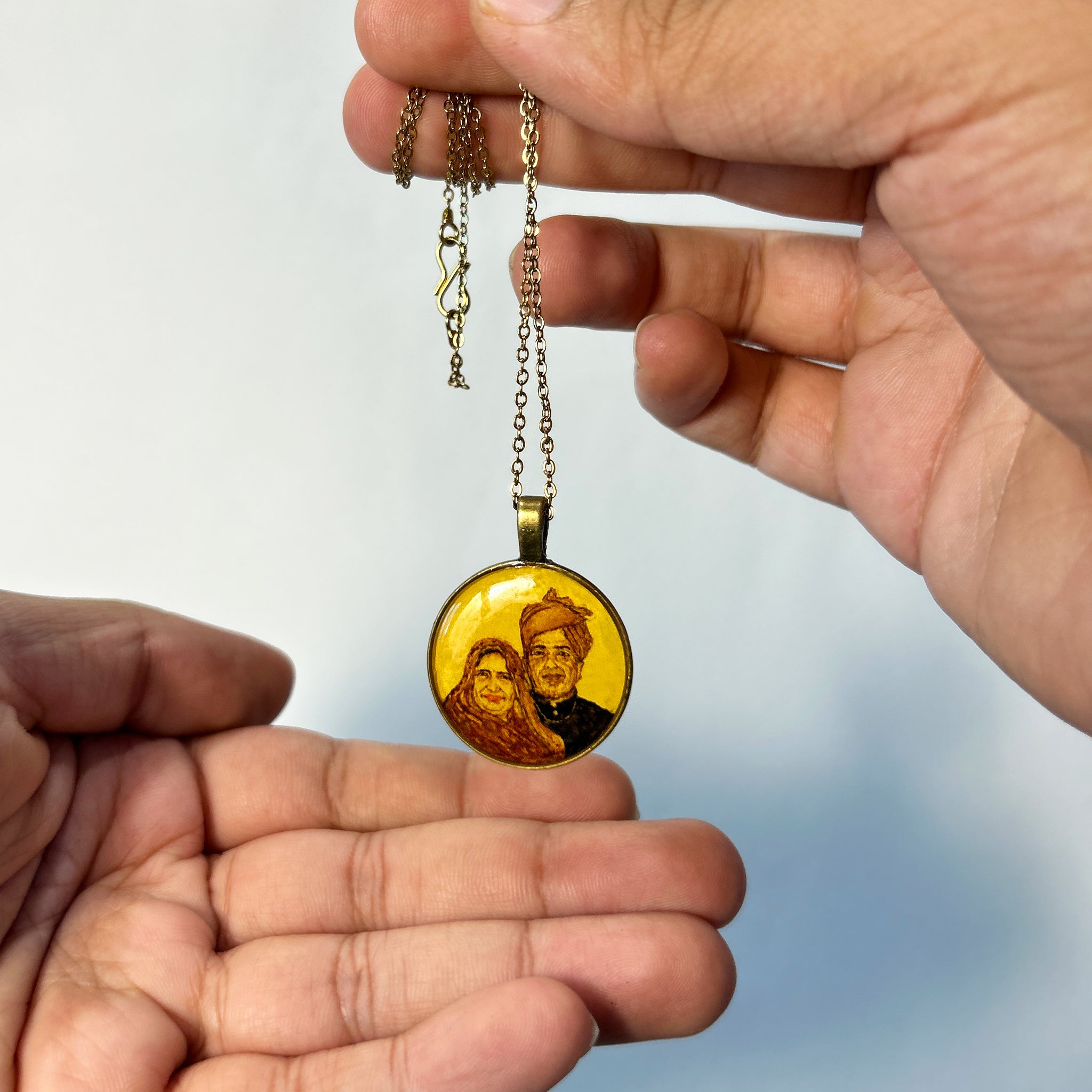 Artistically rendered couple portrait (26mm) capturing traditional Rajasthani essence: husband in pagdi turban and formal attire alongside wife in traditional dress, their warmth evident in gentle smiles. Pendant suspended between hands displaying antique gold colored clasp and chain against serene blue gradient, painted in naples yellow to create heritage appeal. Each cultural detail preserved with luminous protective finish, creating cherished legacy piece.