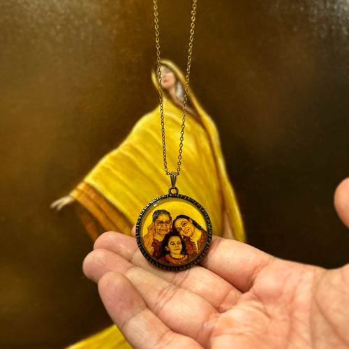 Meticulously rendered heritage portrait (32mm) bridging generations and time: grandmother's gentle presence, mother's loving smile, and precious childhood moment of now-grown daughter, recreated from vintage photograph. Pendant rests in open palm with an antique gold colored chain, photographed against artist's original oil painting where golden sari folds meet deep burnt umber shadows, each detail painted in naples yellow to evoke timeless charm.