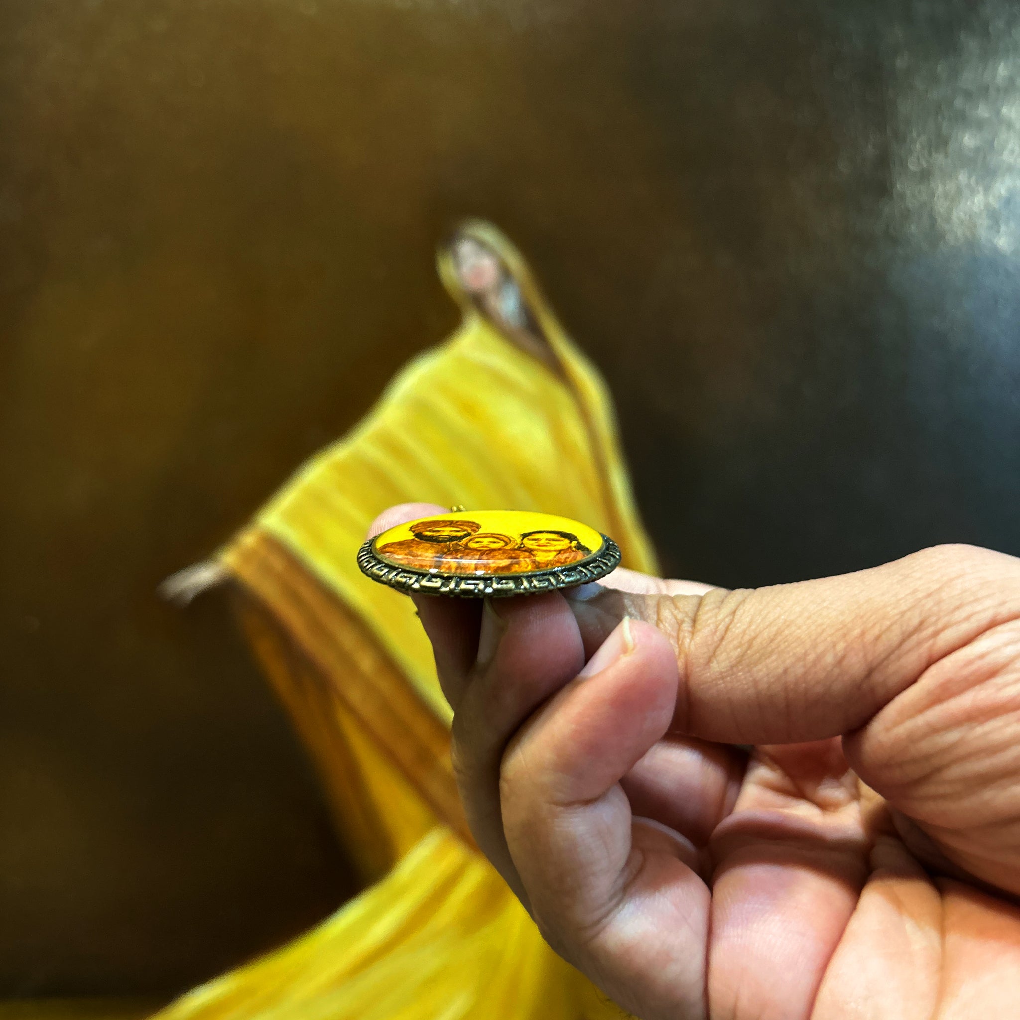 Artistically rendered family portrait (32mm) showcasing dimensional effect: Punjabi family miniature with turban-wearing father, mother, and child, set against naples yellow background with crystal-clear dome coating creating subtle depth. Pendant held horizontally between thumb and forefinger to demonstrate raised surface finish, Greek key border catching light against flowing yellow drapery backdrop. Each brushstroke preserved beneath protective convex sealant in this generational keepsake