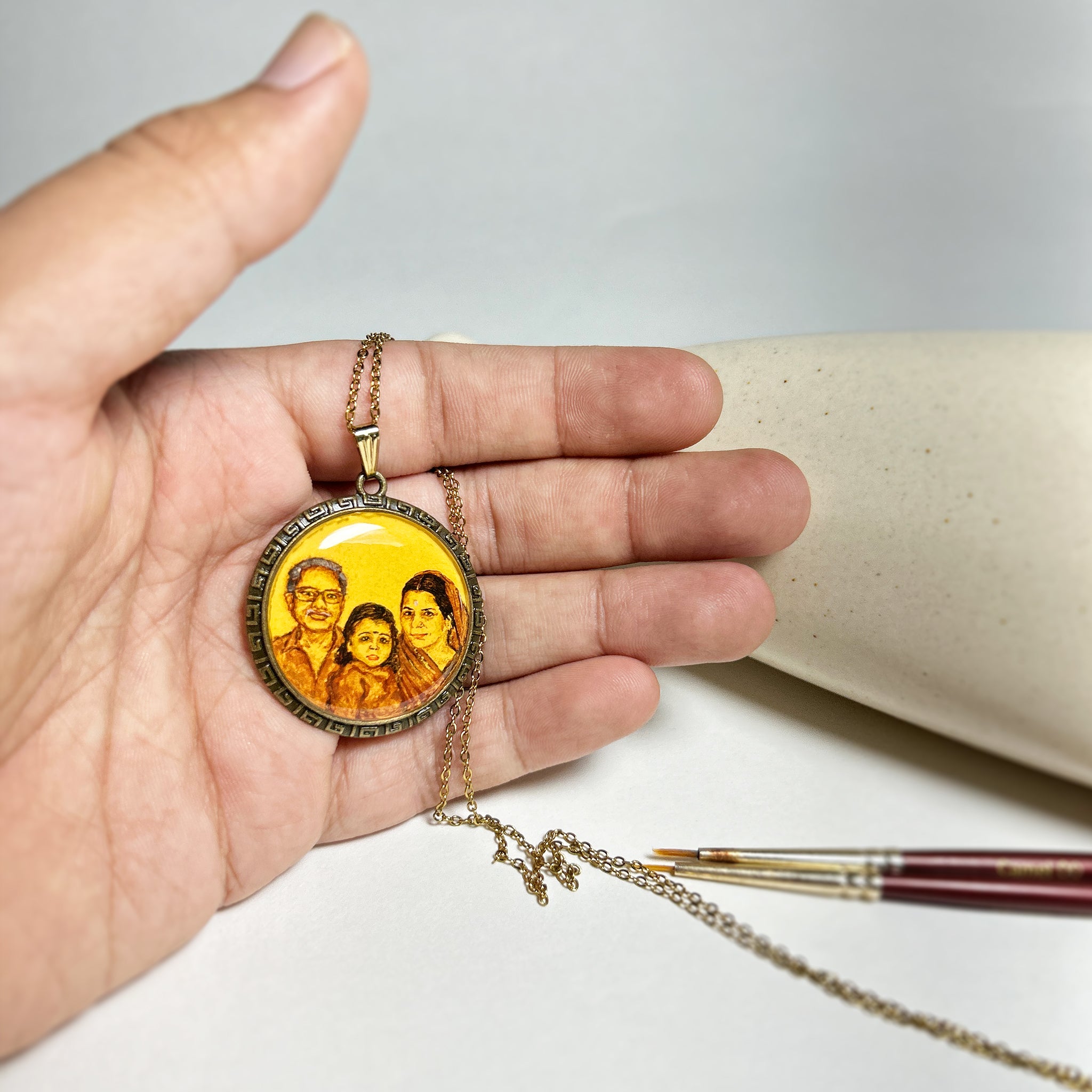 Artistically rendered heritage portrait (32mm) uniting three generations: grandfather's gentle smile behind glasses, grandchild centered with innocent expression, and grandmother's loving gaze, masterfully composed from separate photographs into one harmonious scene. Greek key border frames this petite masterpiece displayed in palm, delicate antique gold finished chain cascading beside artist's brush. Each cherished detail preserved with artisan-grade finish , creating lasting family legacy.