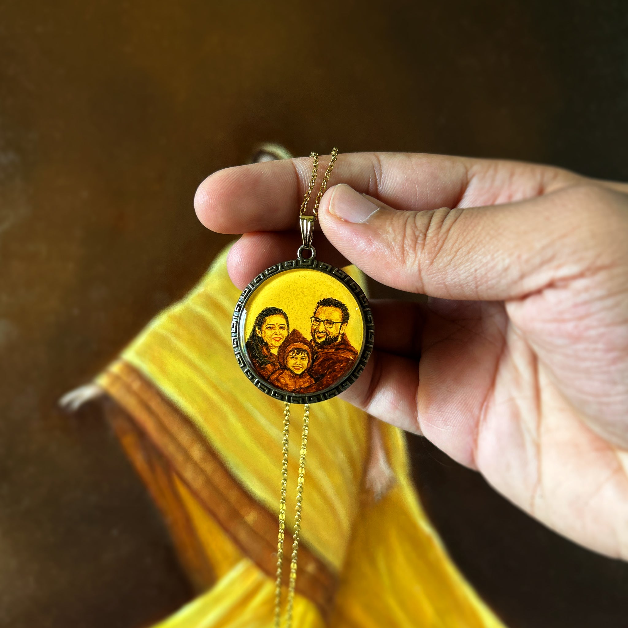 Skillfully crafted diminutive portrait (32mm) capturing intimate family moment: father in glasses, mother's warm smile, and child wearing winter shawl on head, unified in rich sepia tones on naples yellow background. Elegant Greek key pattern frames this treasured memory, pendant held gracefully with delicate antique gold coloured chain  placed between finger and thumbnail against flowing amber backdrop. Each loving detail preserved with museum-grade finish in this enduring family keepsake.