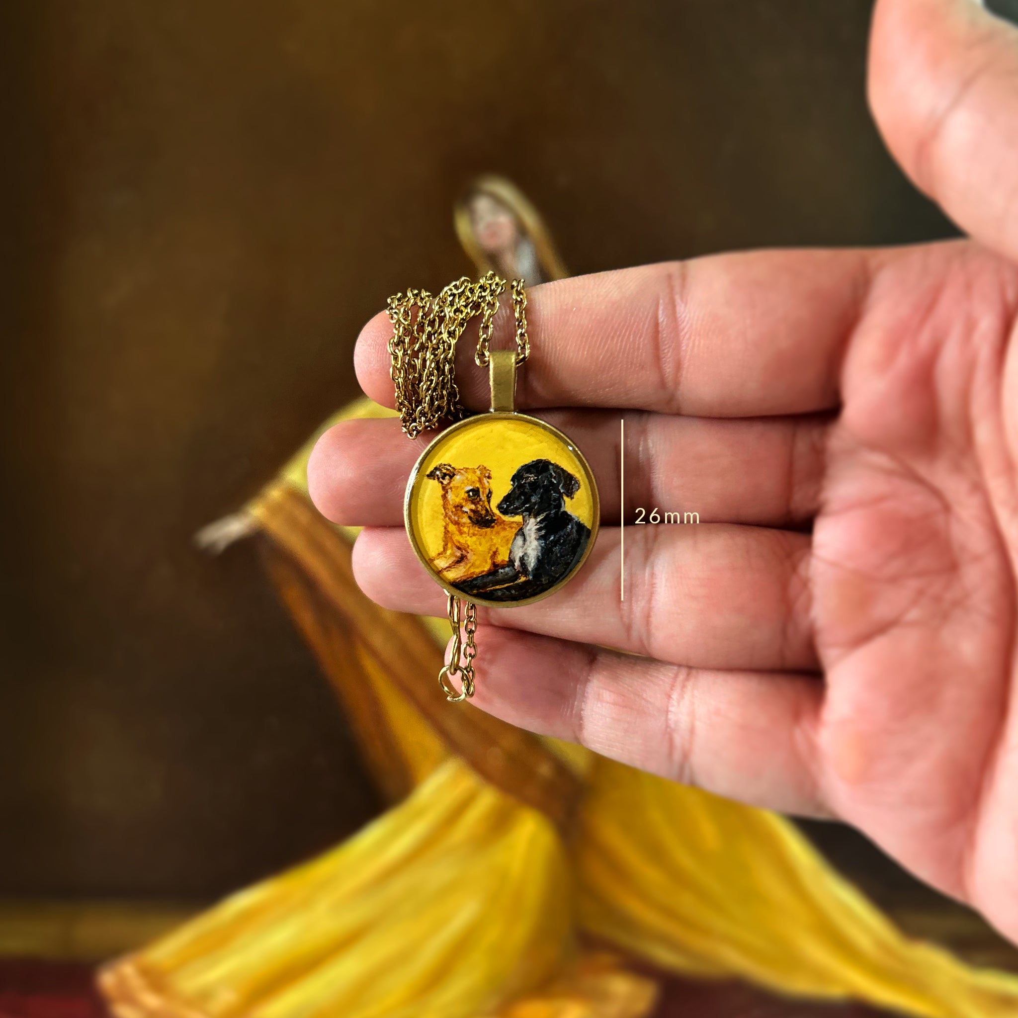 Bespoke pet portrait pendant (26mm) uniting cherished companions: golden-furred and black-coated dogs posed side by side, artistically composed from separate photos into one heartwarming scene on naples yellow background. Pendant displayed in open palm with measurement guide, delicate chain cascading over fingers, against artist's original oil painting with dramatic yellow drapery, enhanced with luminous protective layer to preserve this unique dual memorial keepsake.