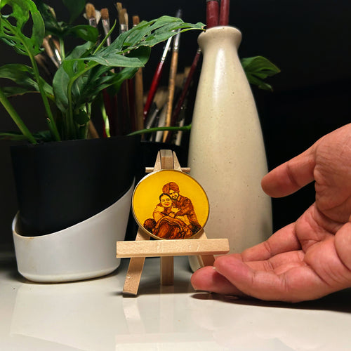 Hand-painted miniature (5cm) immortalizing tender romance: Sikh husband in pagadi draws wife into full embrace, his arms encircling her shoulders while she leans into his protective hold, their radiant smiles reflecting deep affection. Artwork nestled between black-white planter with cascading monstera leaves and ceramic vase, paintbrushes visible in background, all enhancing this vintage-styled portrait on naples yellow canvas amidst modern home decor,where intimate expressions span smaller than thumbnail.