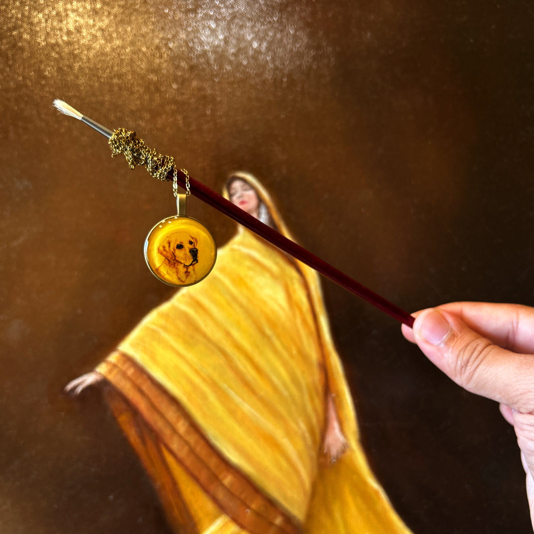 Artistically styled photograph: Labrador portrait pendant (2.6cm) suspended from red-handled paintbrush, chain elegantly draped against ethereal golden-draped backdrop. Miniature captures beloved companion's gentle expression and loyal eyes in warm sepia tones on naples yellow background, classic round frame enhanced with luminous protective layer. Hand holding brush creates dramatic diagonal composition, demonstrating unique display of this heirloom-quality memorial piece.
