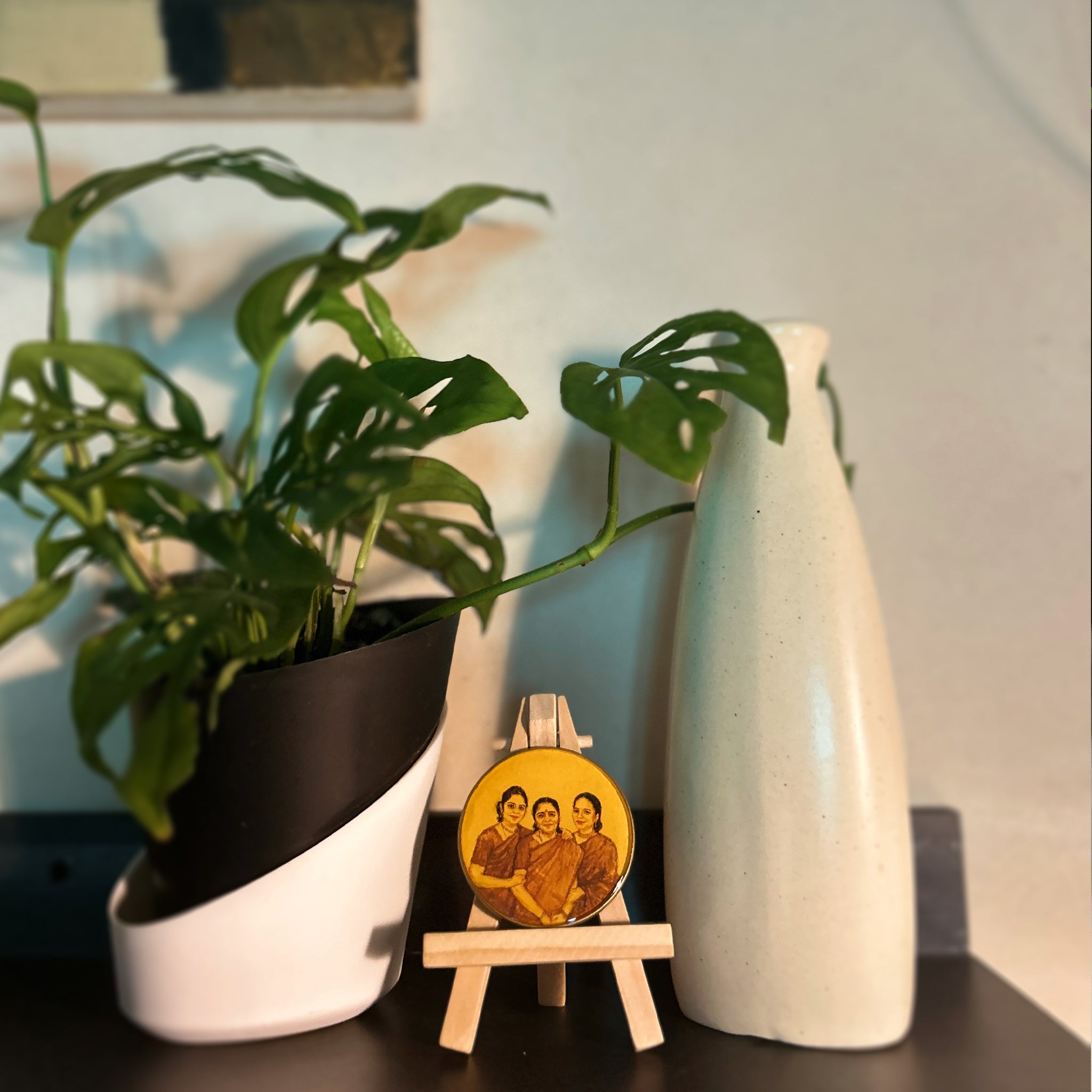 Hand-painted miniature (5cm) featuring harmonious family portrait: mother centered between daughters, all in traditional saris sharing warm smiles and gentle embrace, painted in rich brown and burnt umber on naples yellow background. Wide-angle capture reveals artwork's delicate scale on wooden easel, nestled between oversized monstera planter and tall ceramic vase, demonstrating how this vintage-styled, heirloom-quality piece creates intimate focal point amid contemporary home decor.